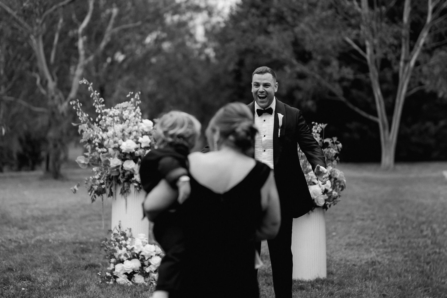 Black and white wedding photo showing guests and floral arrangements during an outdoor ceremony.