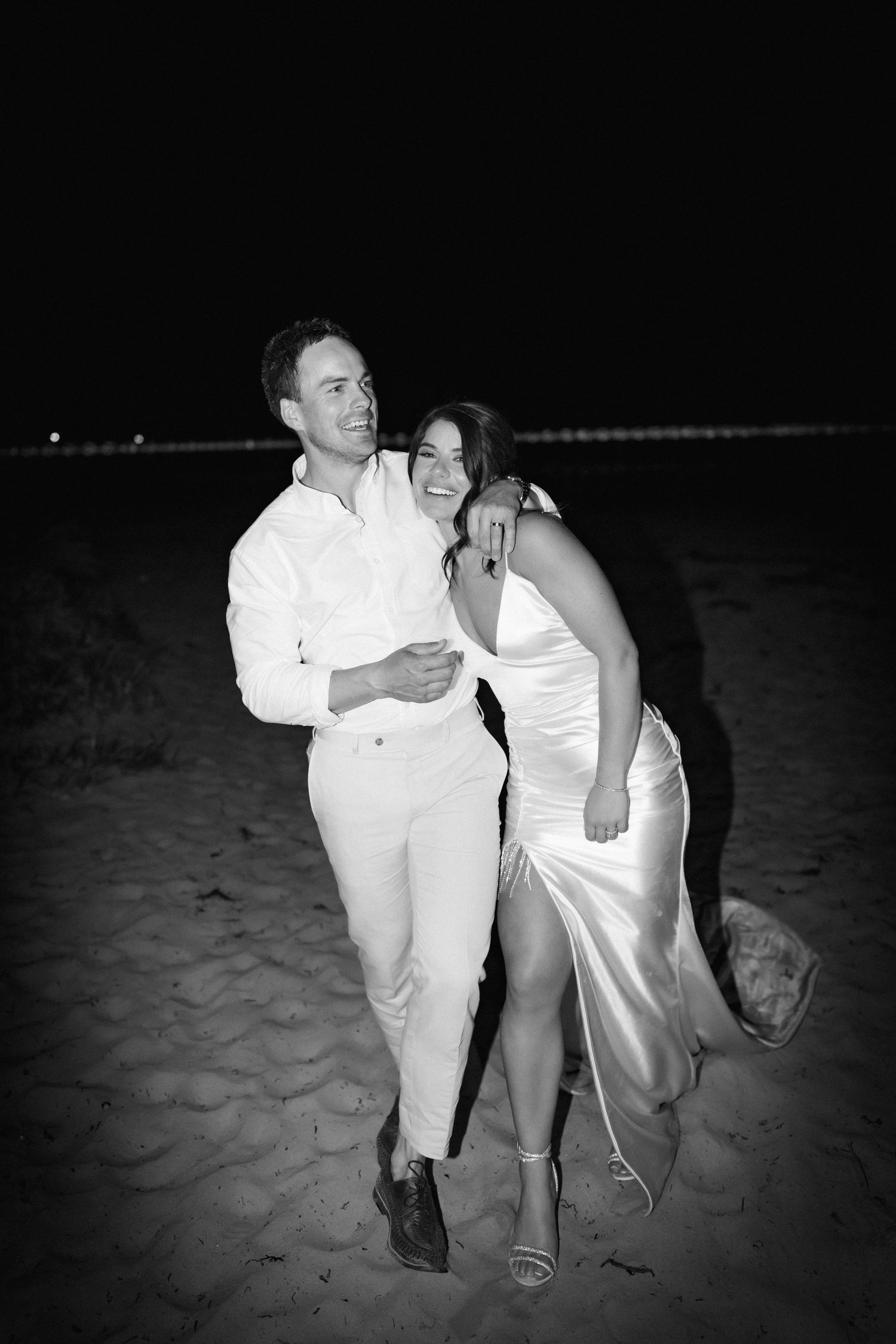 A couple shares a joyful moment on the beach at night in matching white outfits.