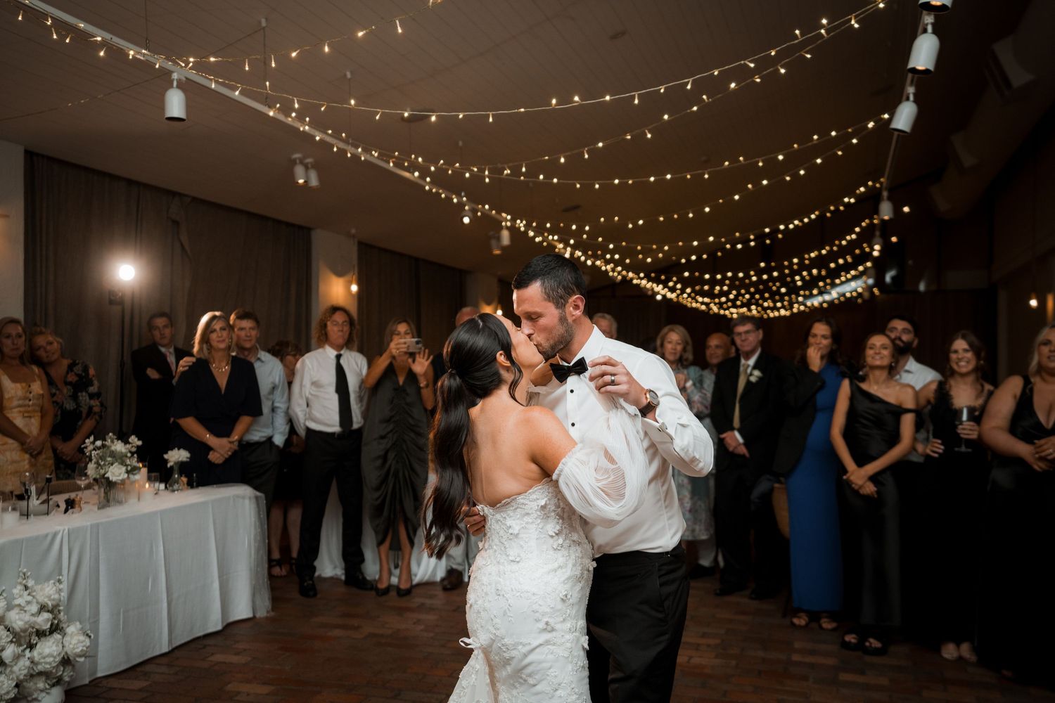 Newlyweds share their first dance under twinkling string lights at their reception venue.