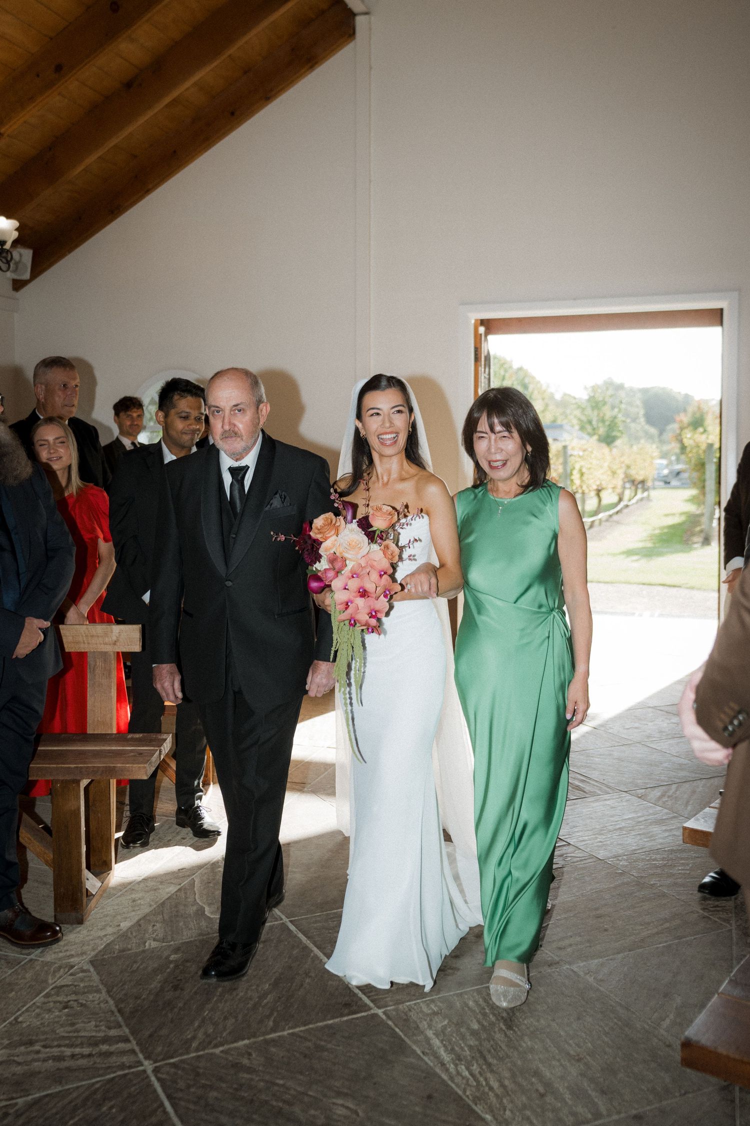 Bride walks down the aisle escorted by parents in a rustic chapel, holding a colorful bouquet.