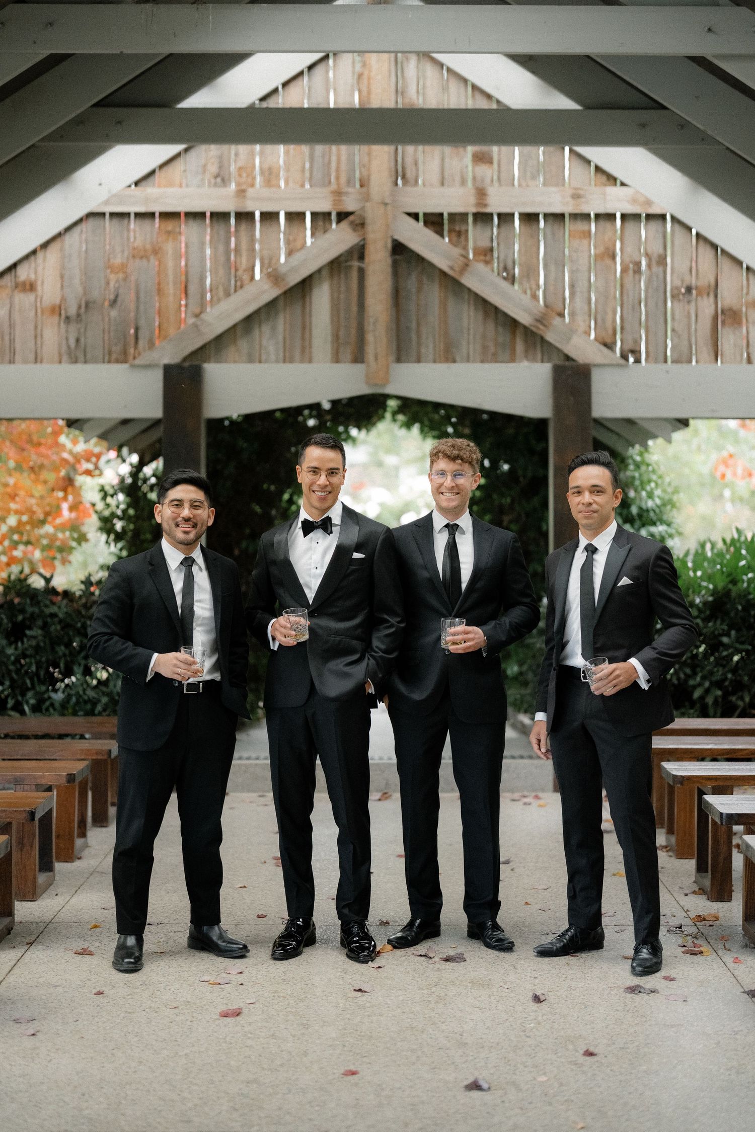 Four groomsmen in black tuxedos holding drinks pose together at an outdoor wedding venue with wooden chapel architecture.