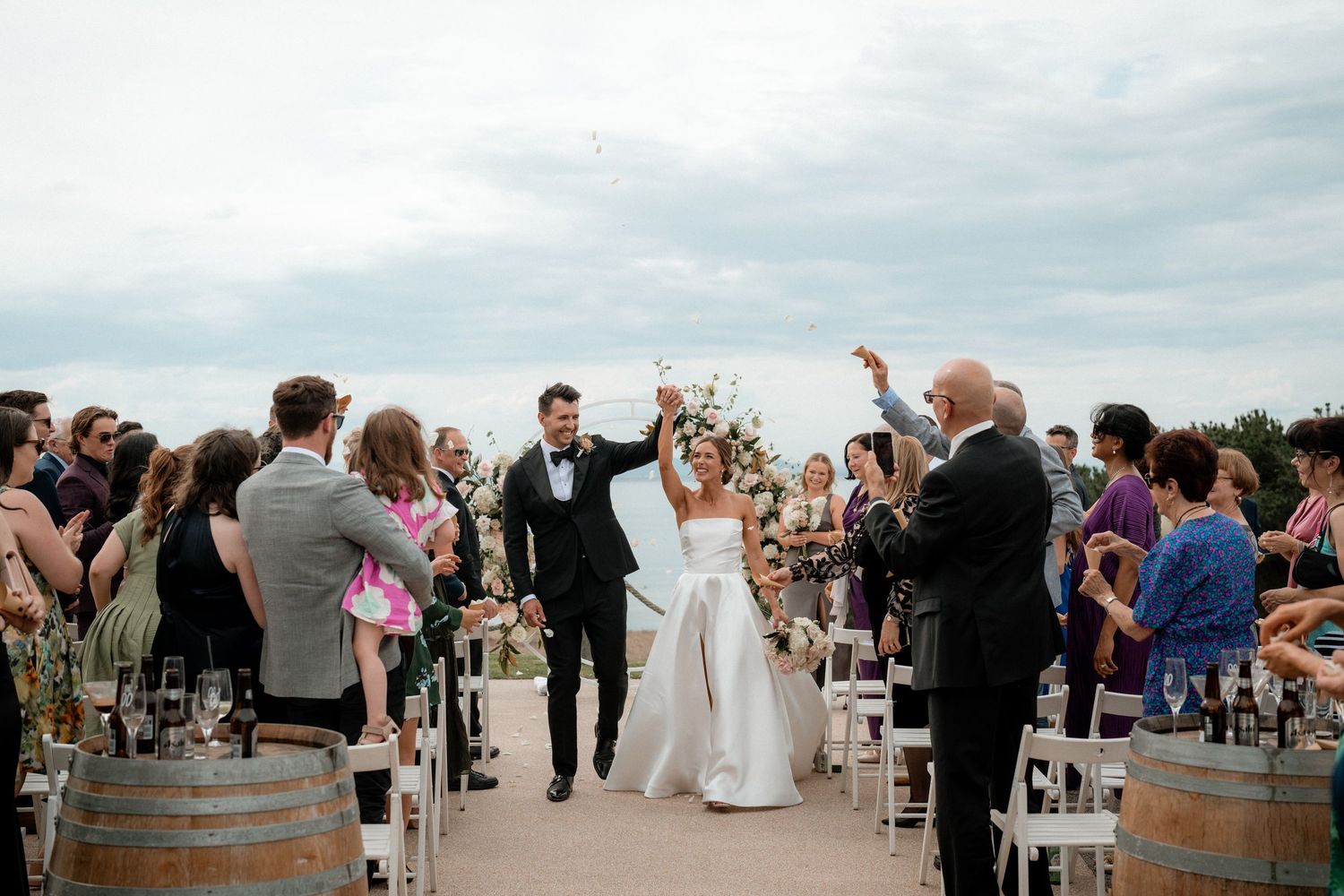 A panoramic sequence of outdoor wedding ceremony moments with guests gathered between rustic wine barrels at sunset.