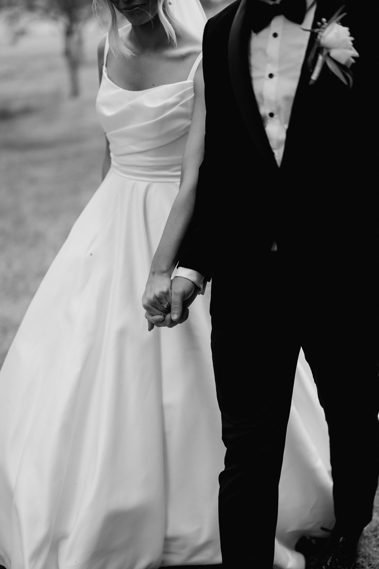 Black and white close-up of bride and groom holding hands, showing elegant wedding dress and tuxedo details.
