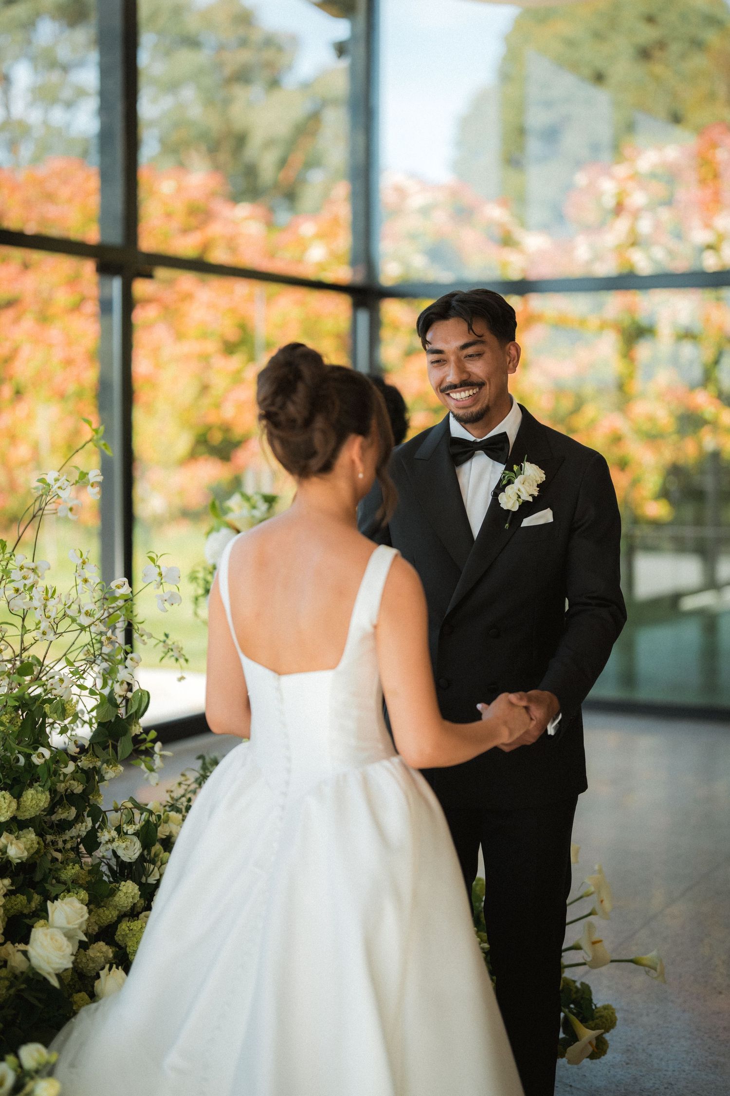 A groom in a black tuxedo and bow tie smiles while holding hands with his bride in a white gown beside white floral arrangements.
