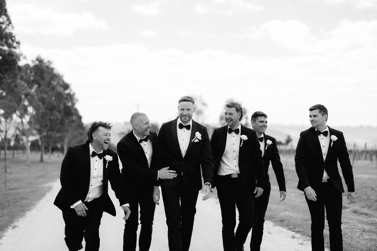 A group in black tuxedos walking and laughing together in a candid black and white photograph.