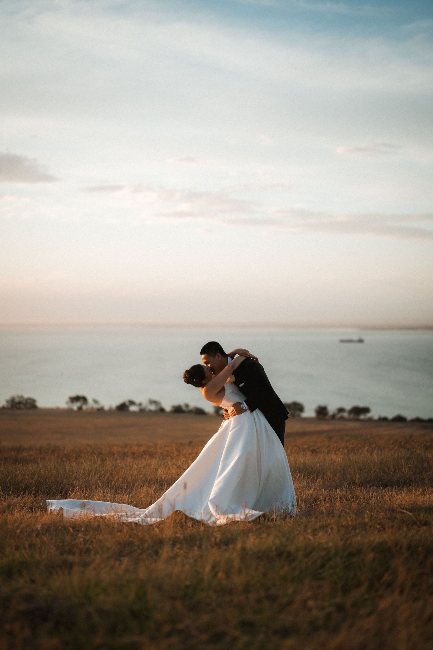 A romantic sunset wedding photo in a golden field with the ocean visible in the distance.
