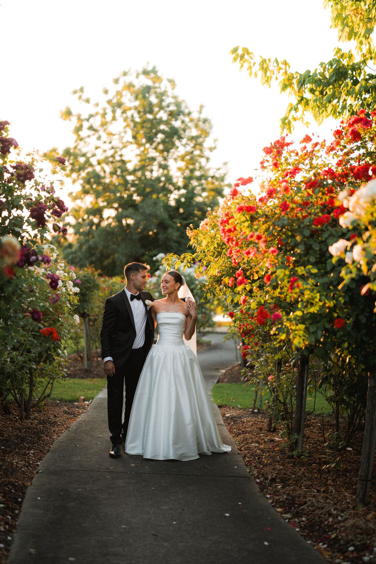 A couple walks along a garden path with vibrant red flowers and autumn foliage creating a romantic setting.