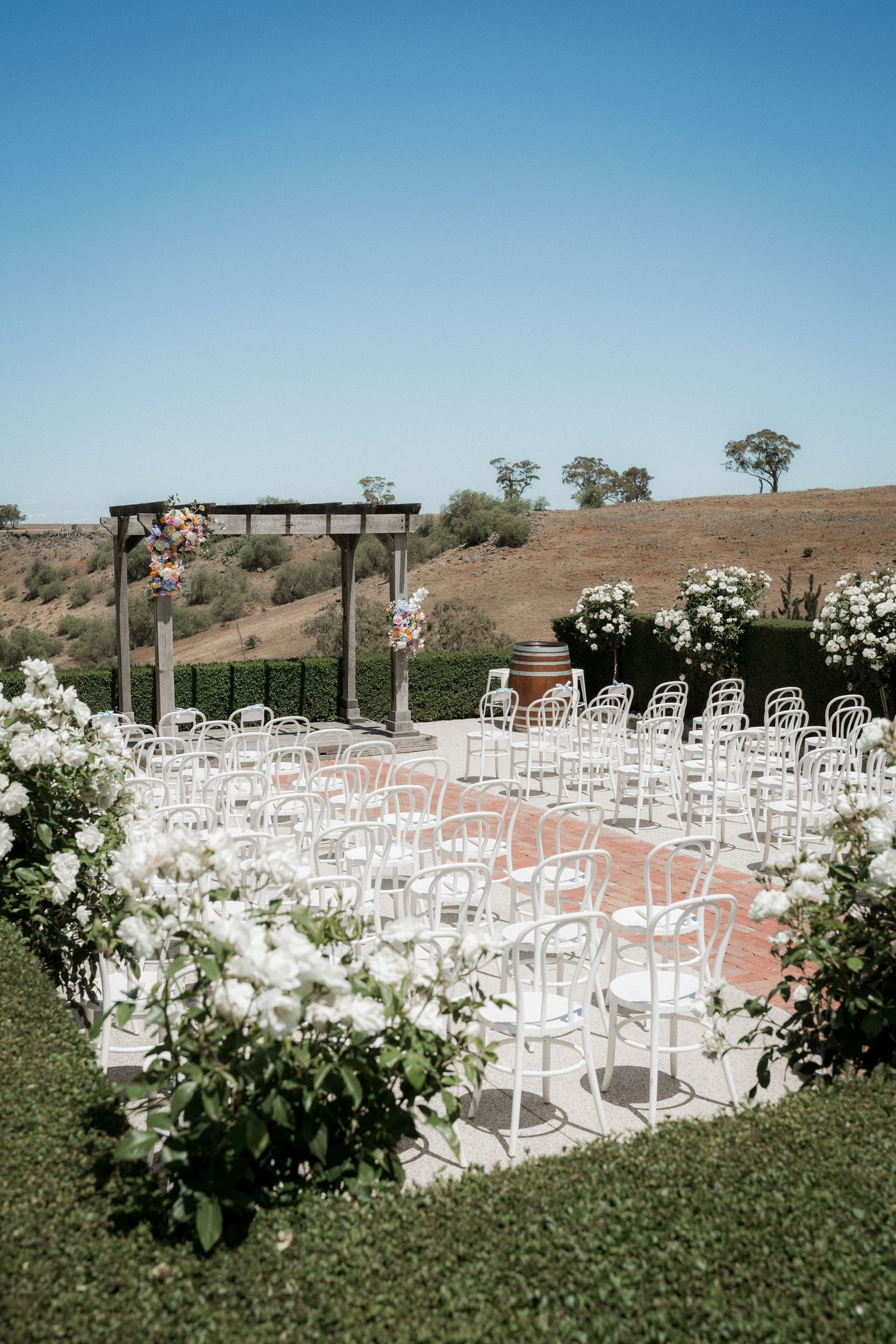 Elegant outdoor wedding ceremony setup with white chairs, wooden arch, and rolling hills backdrop under blue sky.