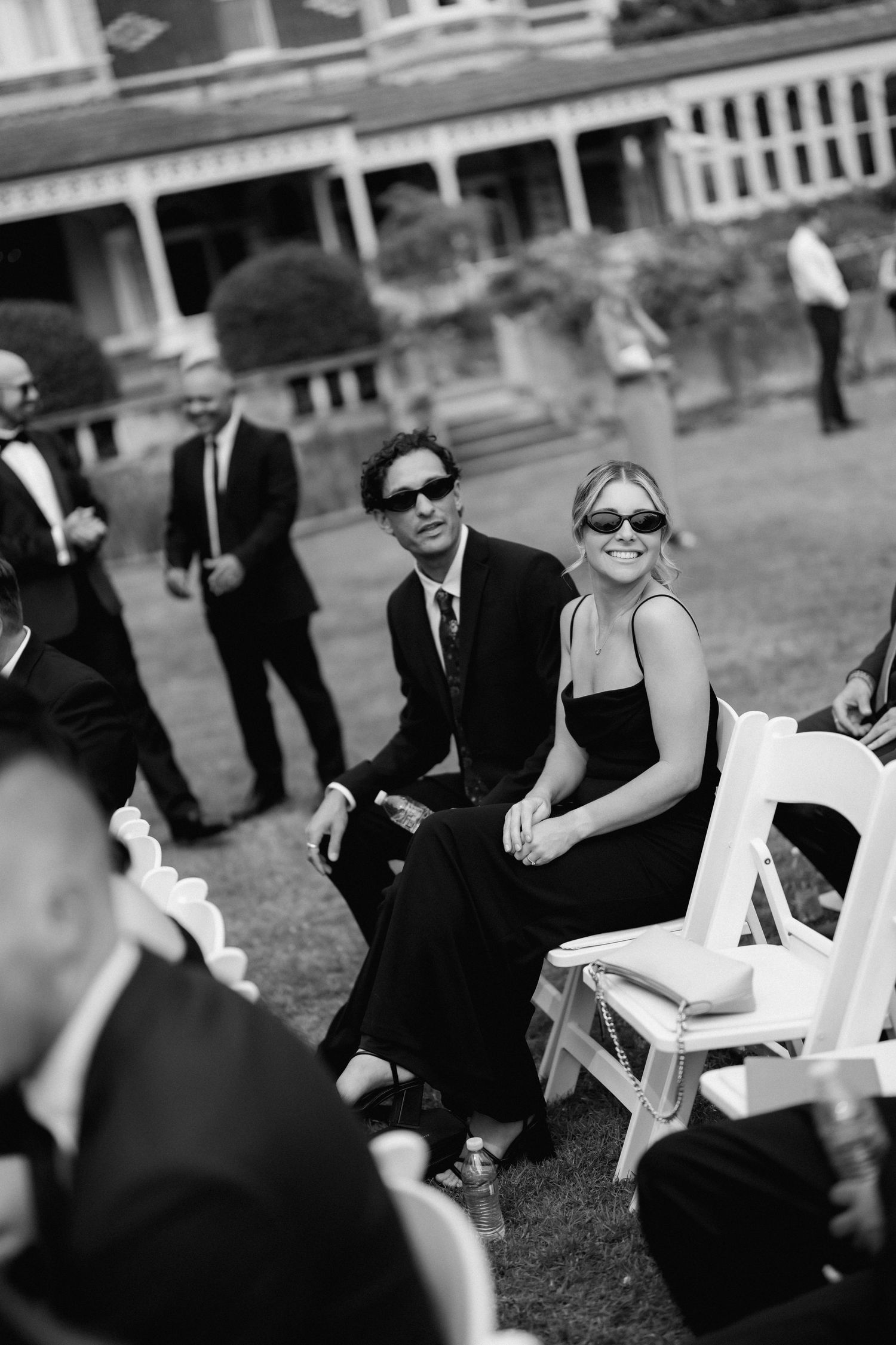 Black and white photo of elegantly dressed guests in sunglasses seated at an outdoor event.