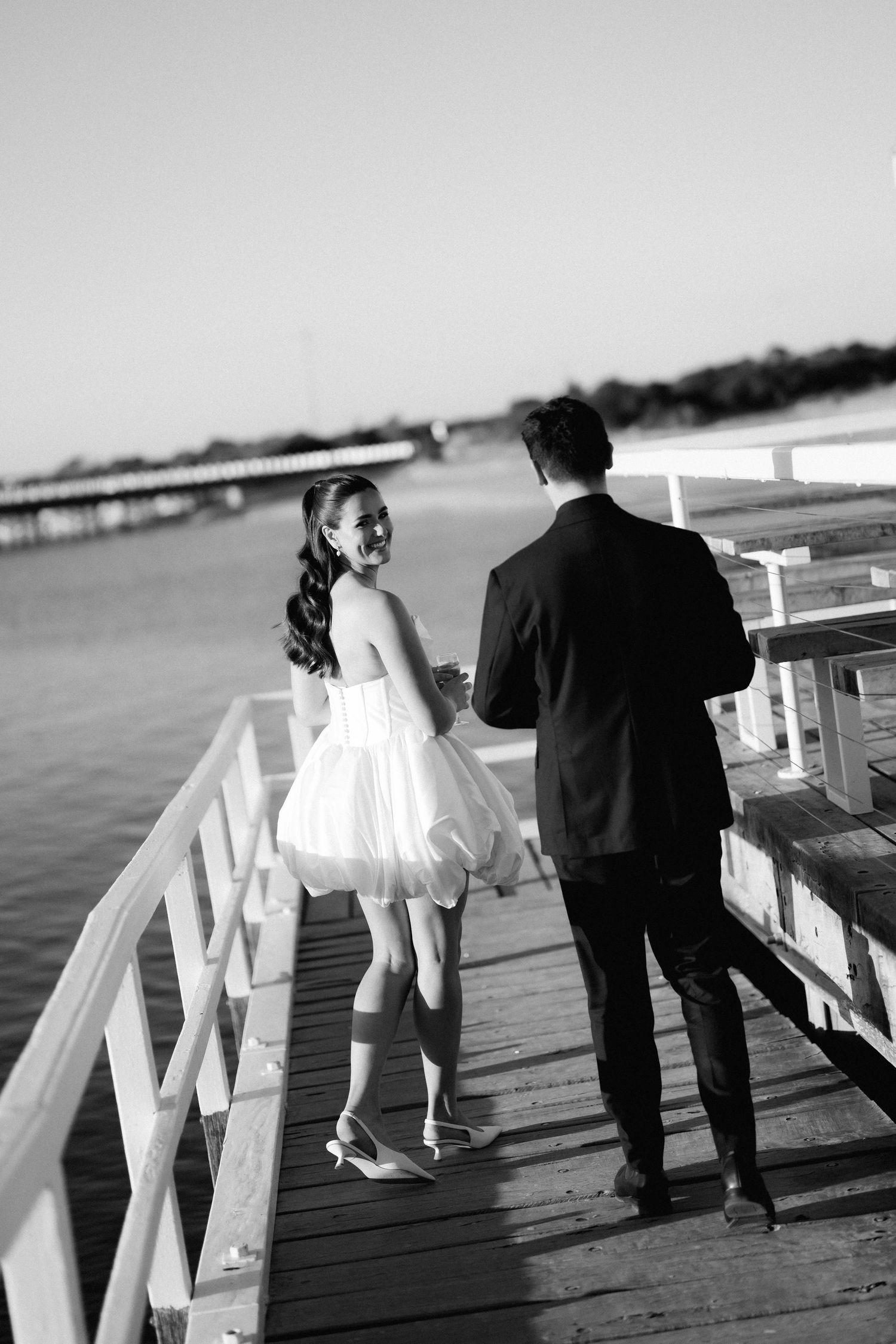 A couple shares a romantic moment on a white wooden pier overlooking the water on a sunny day.