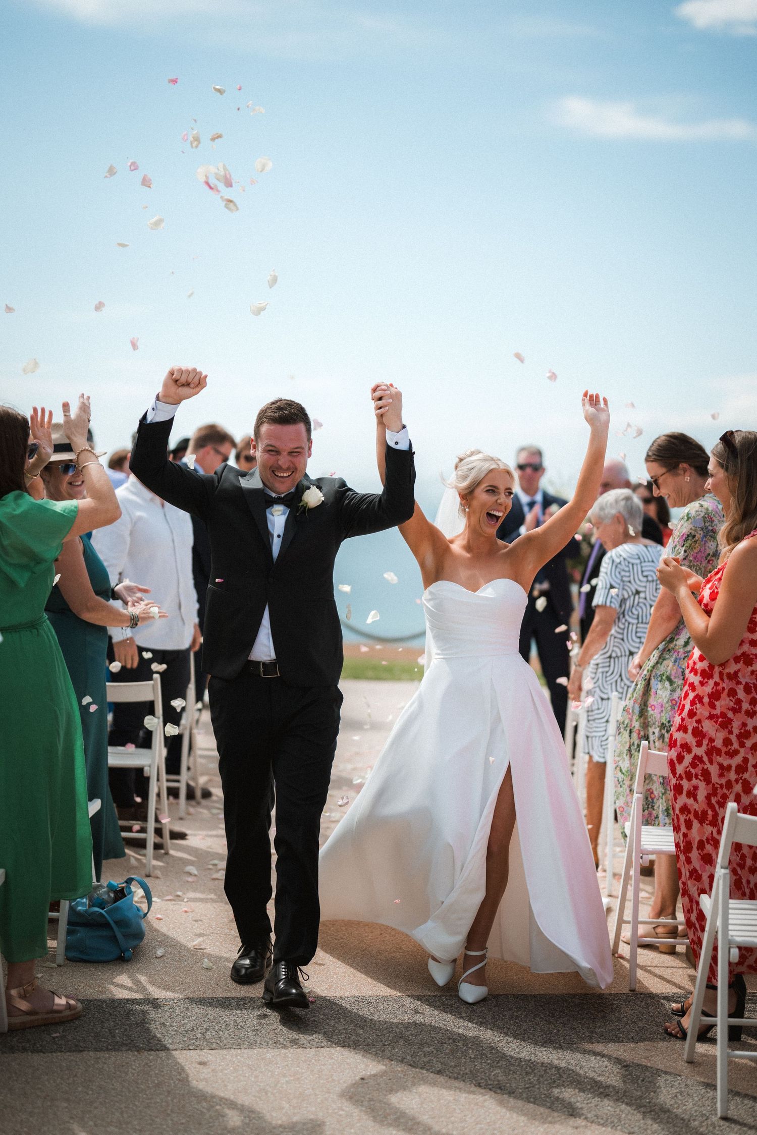 Joyful wedding celebration with confetti toss on a sunny coastal pathway.