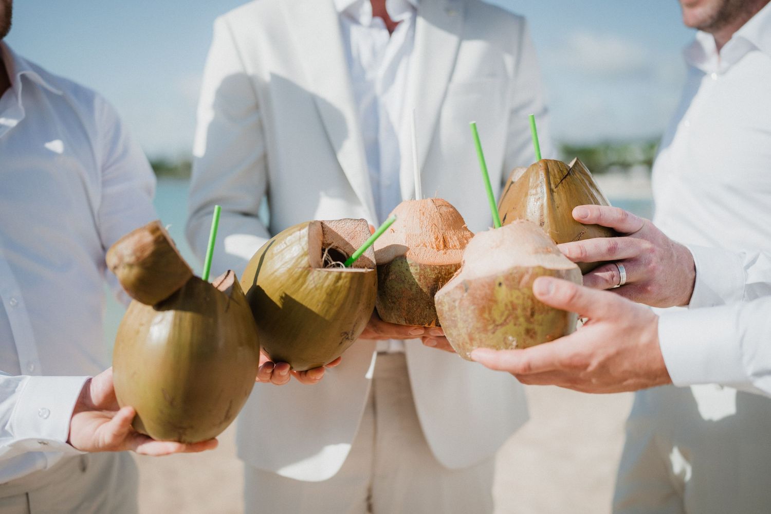 Fresh coconuts with straws being served at a tropical beach wedding reception with people in white attire.