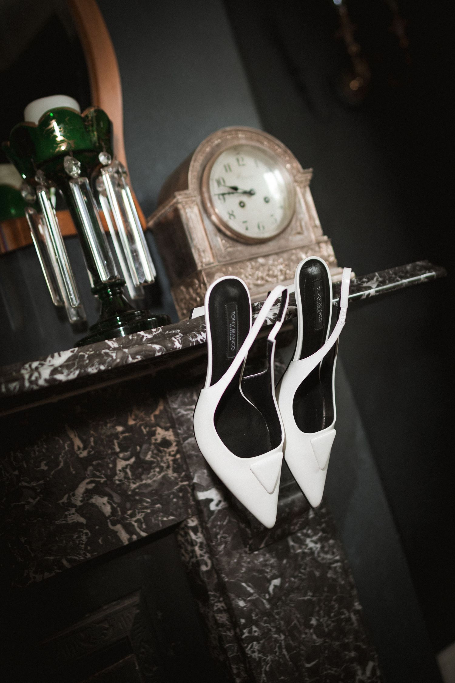 Elegant white wedding heels displayed next to a vintage clock and green glass bottle on a marble surface.