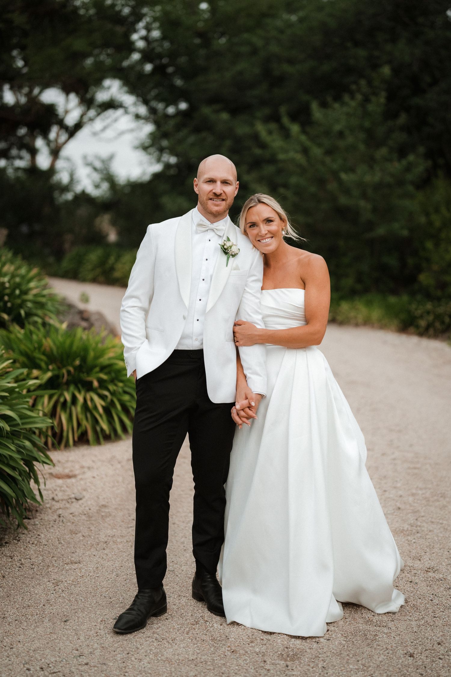 A couple in wedding attire poses on a garden path with greenery in the background.