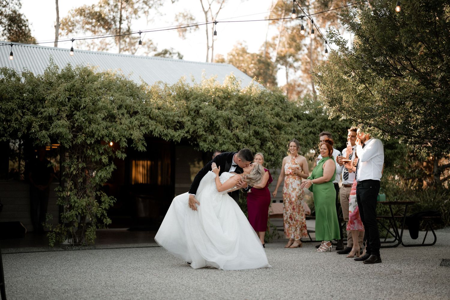 A wedding party poses together outdoors on a paved area surrounded by lush greenery at dusk.