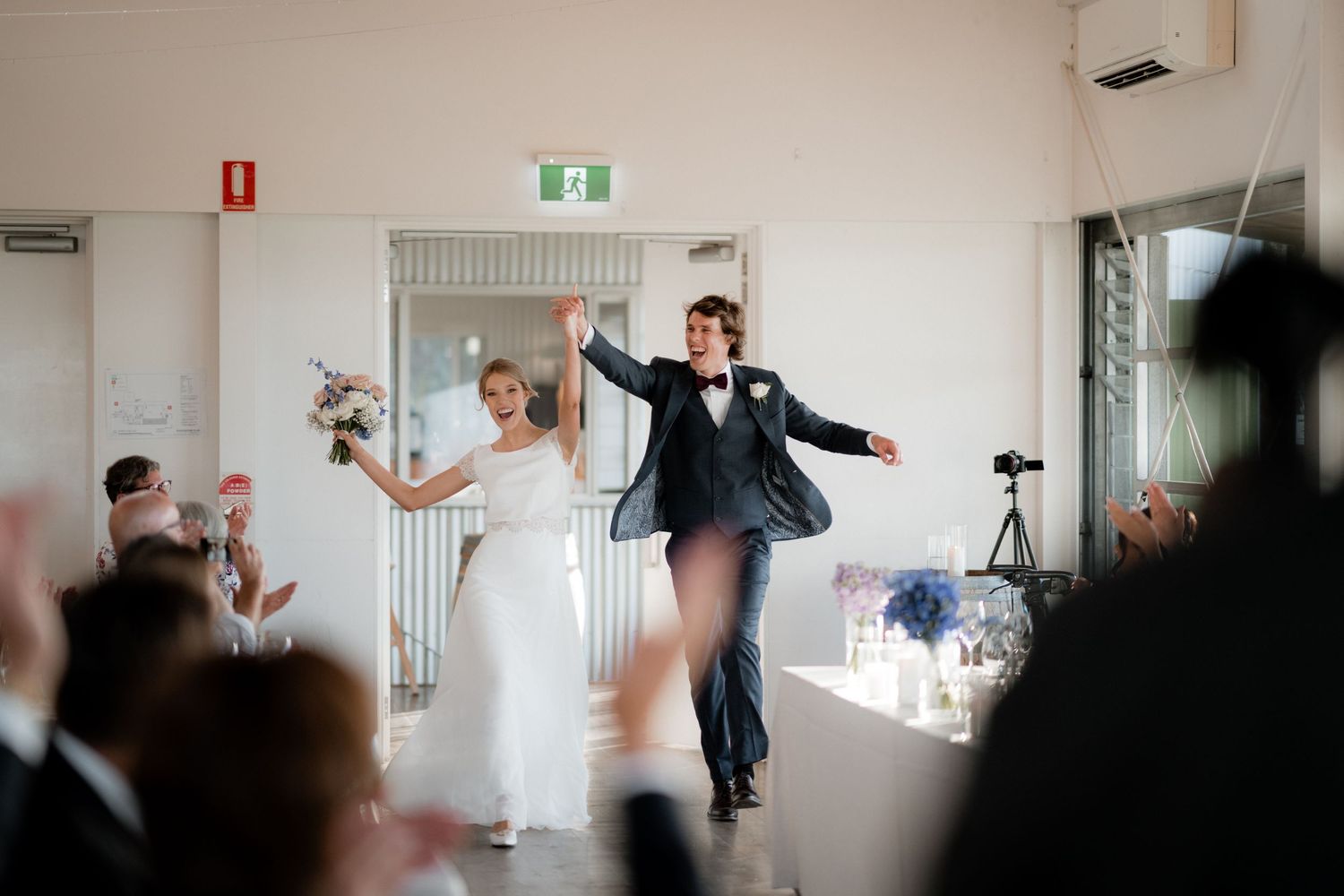 A joyful wedding couple triumphantly makes their entrance into a bright reception venue with guests cheering.