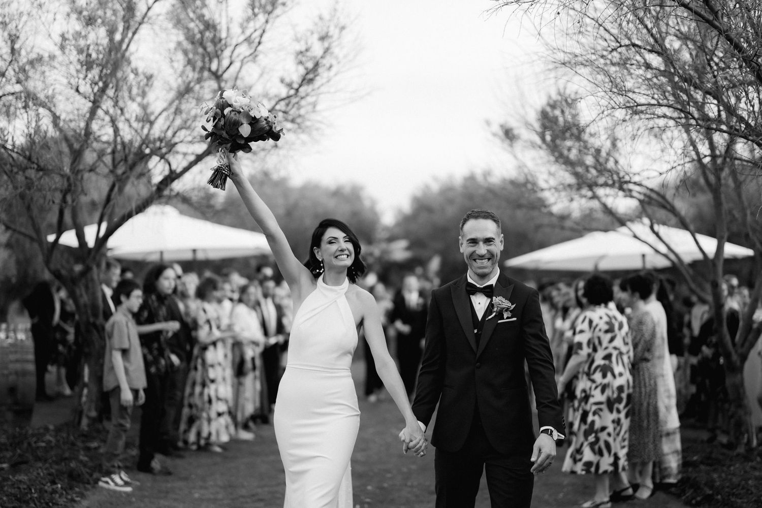 Happy newlyweds celebrate during outdoor wedding reception while holding hands and raising bouquet in black and white.