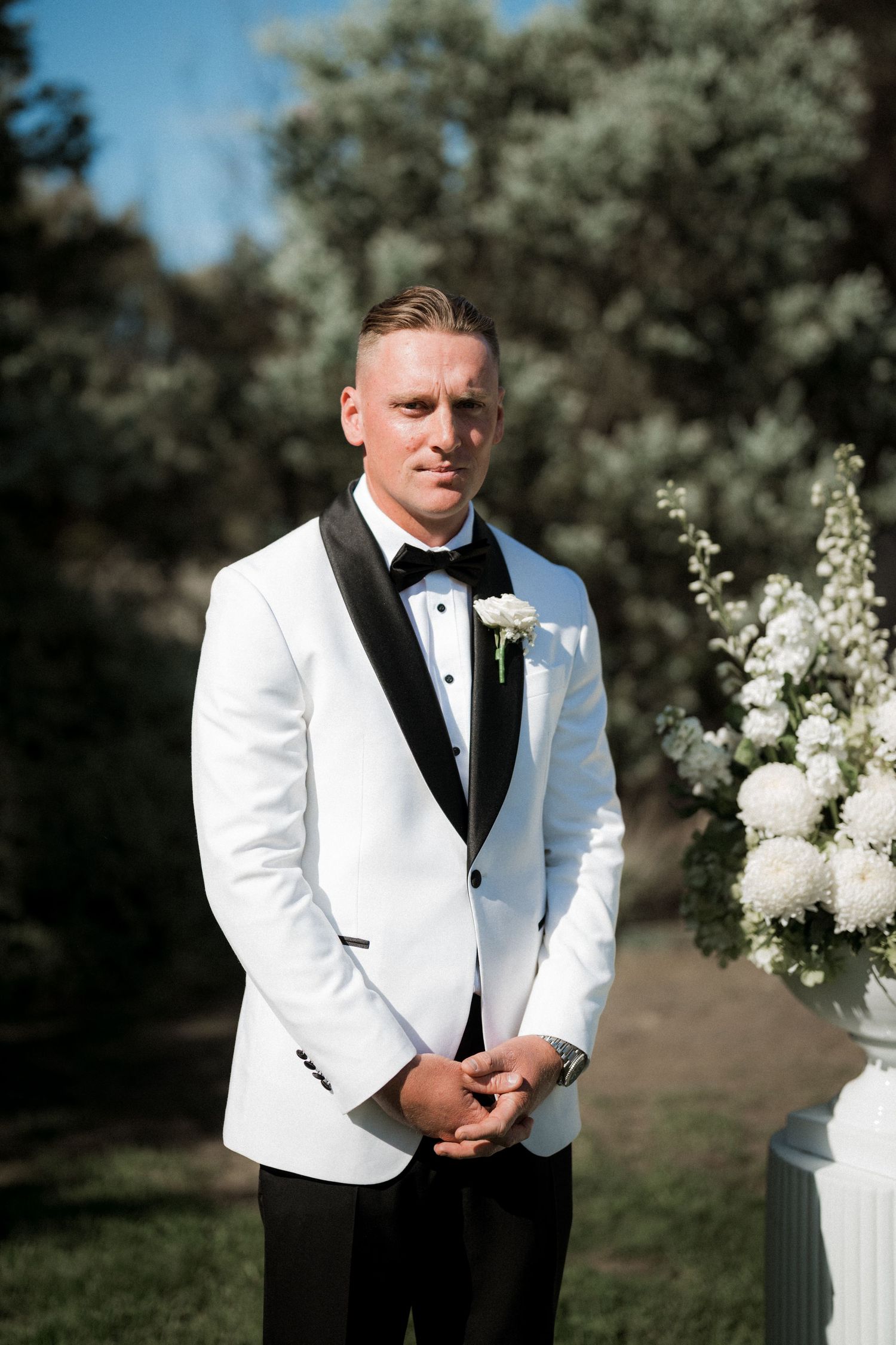 A groom in a white tuxedo jacket and black bow tie poses for formal wedding portraits in an outdoor garden setting.