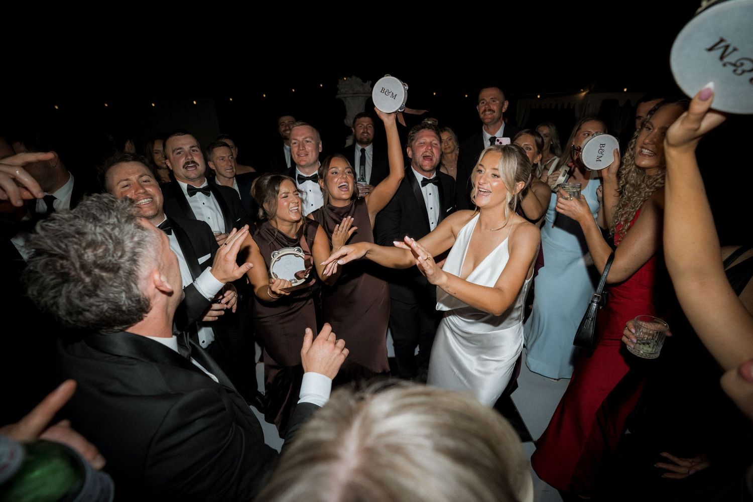 Wedding guests dance and celebrate on a dimly lit dance floor at an evening reception party.