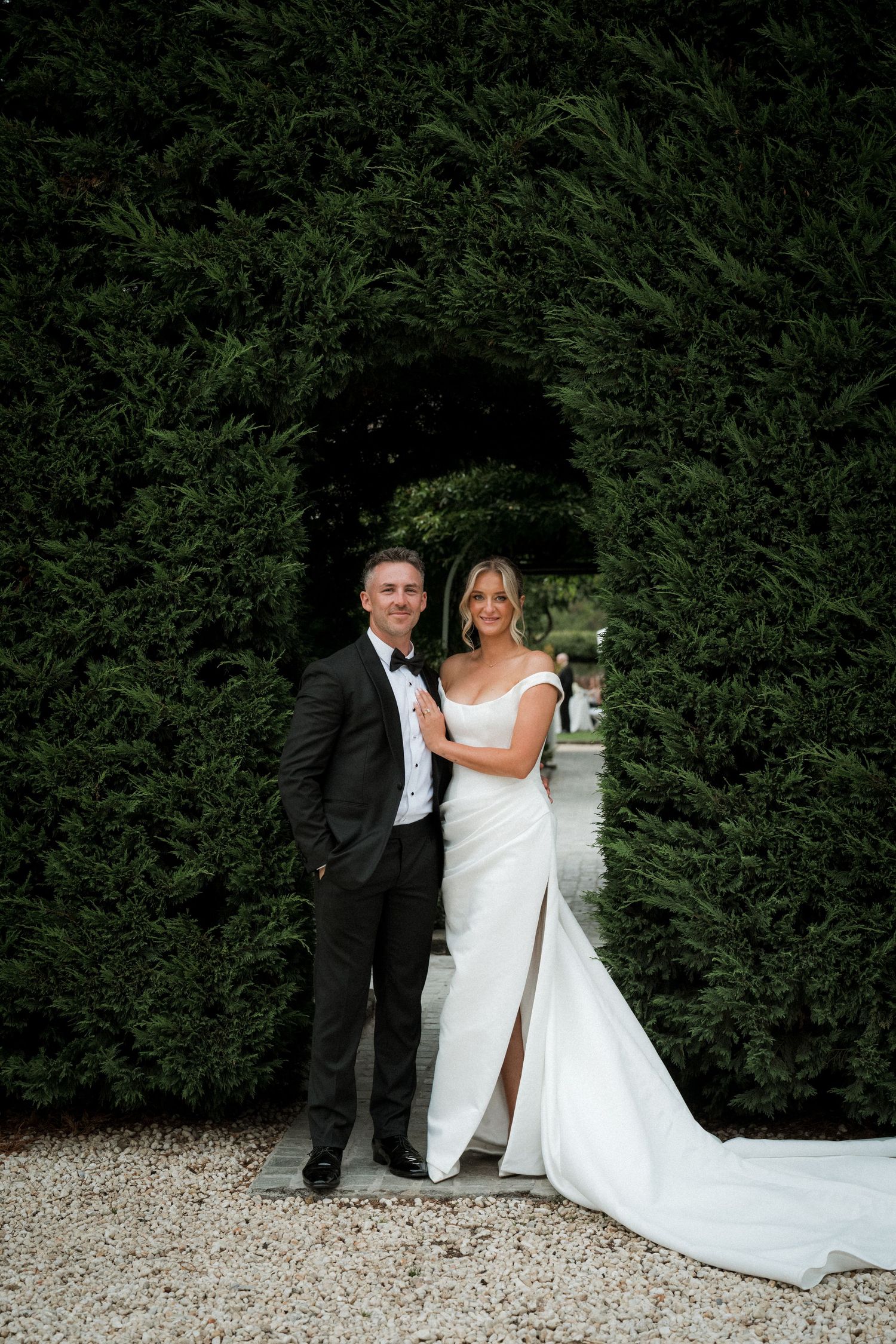 Wedding couple poses against hedge backdrop in formal attire.