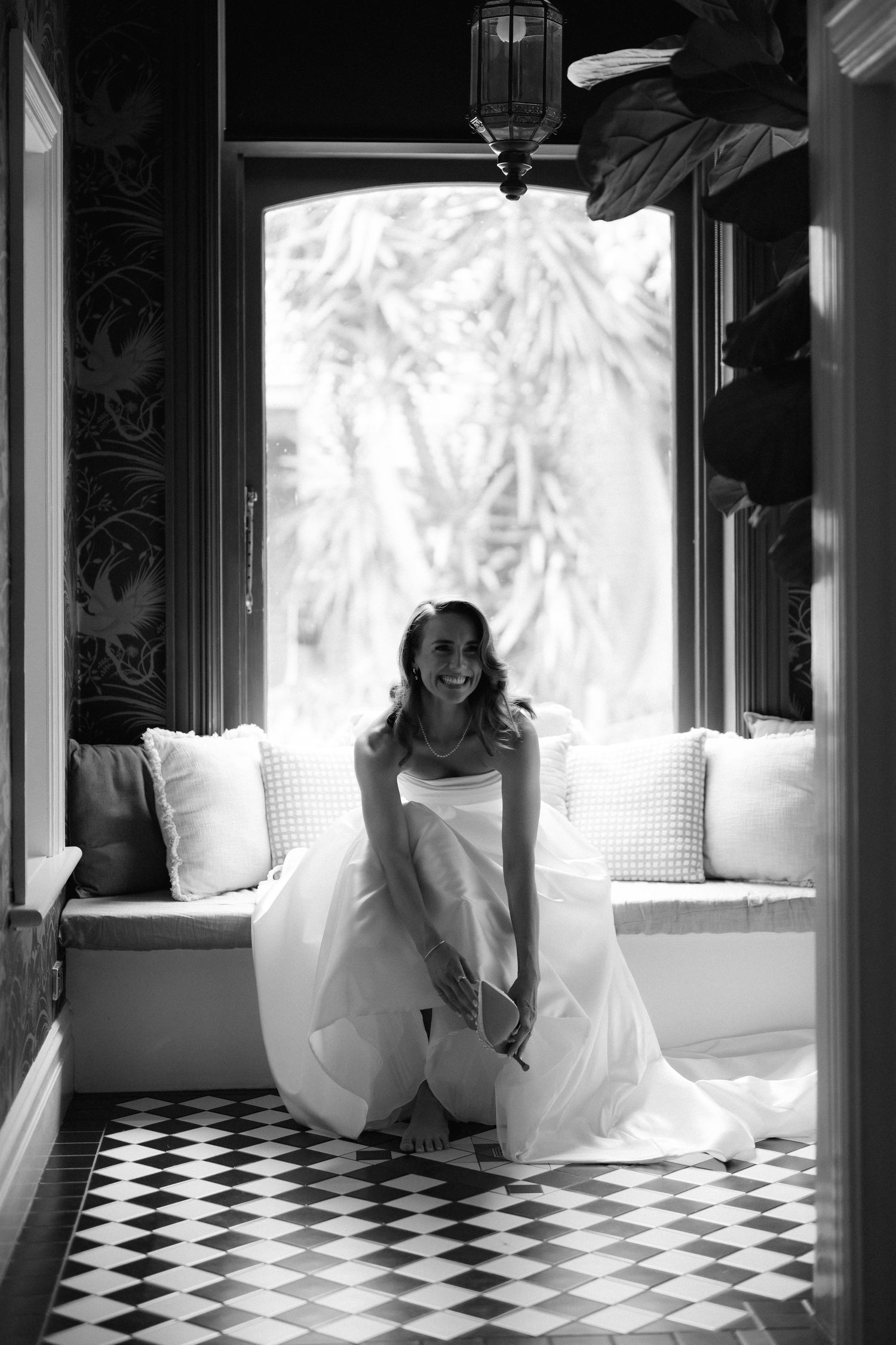 Bride in white dress sitting on checkered floor in front of bright windows.