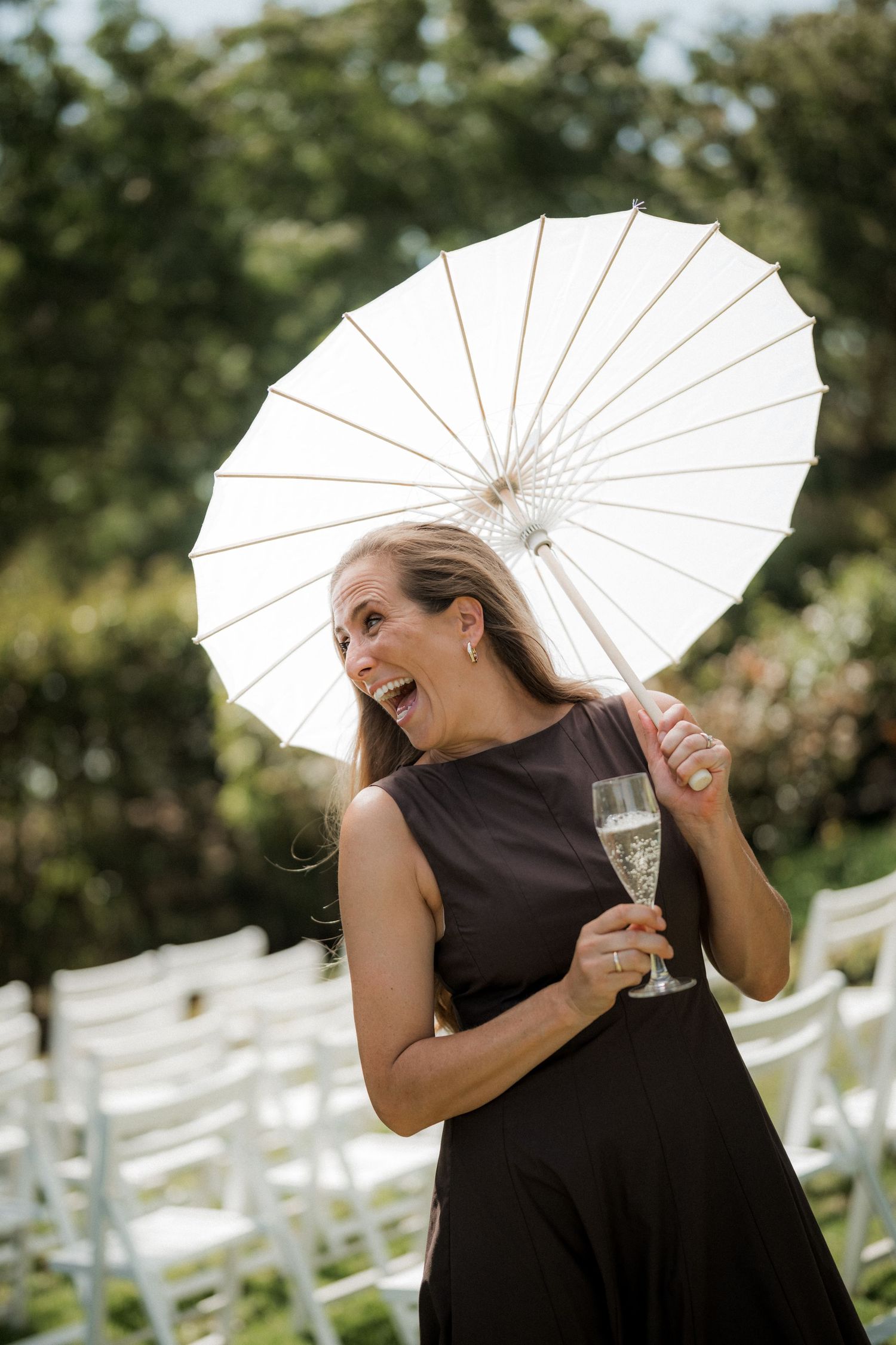 A guest in a black dress holds a white parasol and champagne glass while laughing at an outdoor wedding.