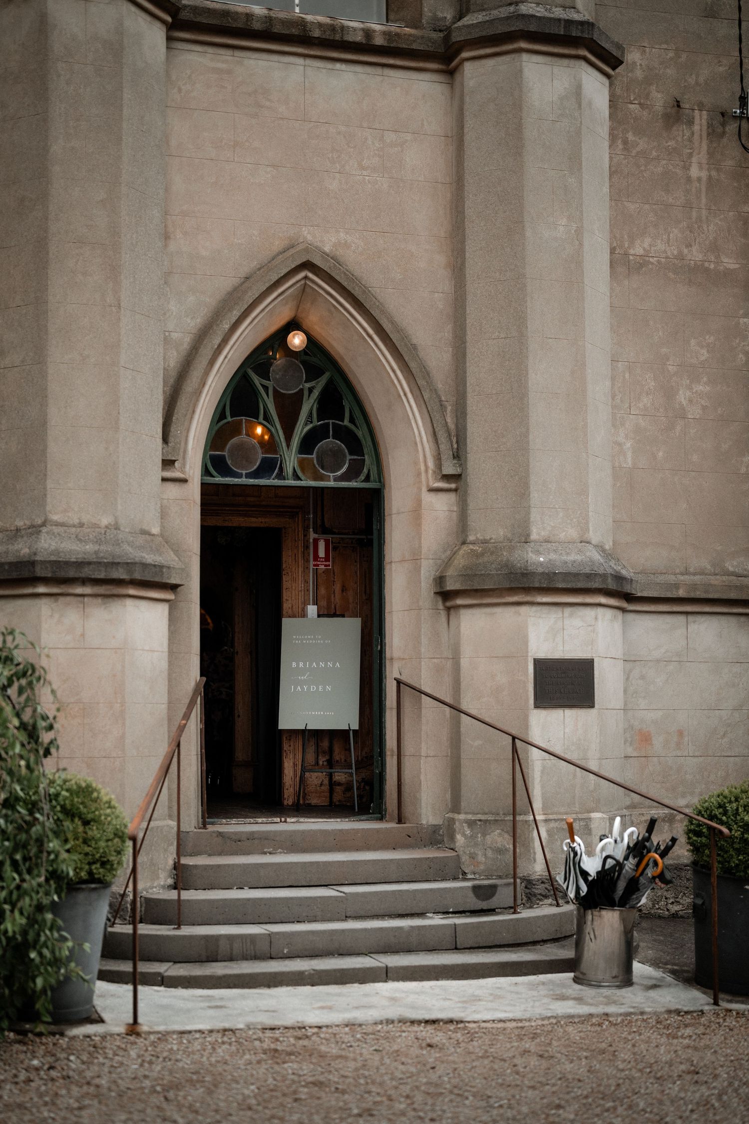 Gothic stone church entrance with arched doorway, stone steps, handrails, and potted plants framing the entrance.