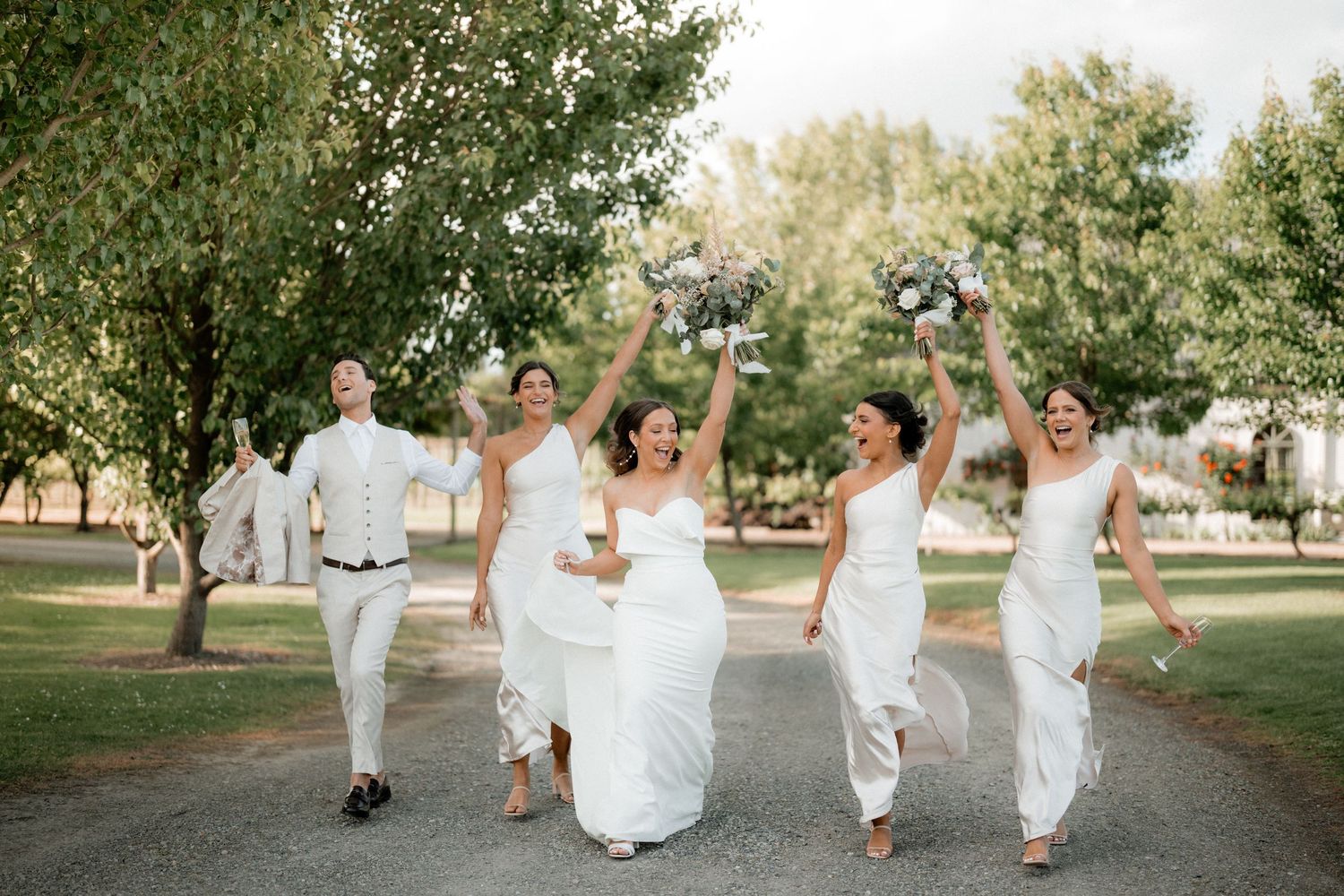 A wedding party in white attire walking and celebrating joyfully down a tree-lined path on a sunny day.