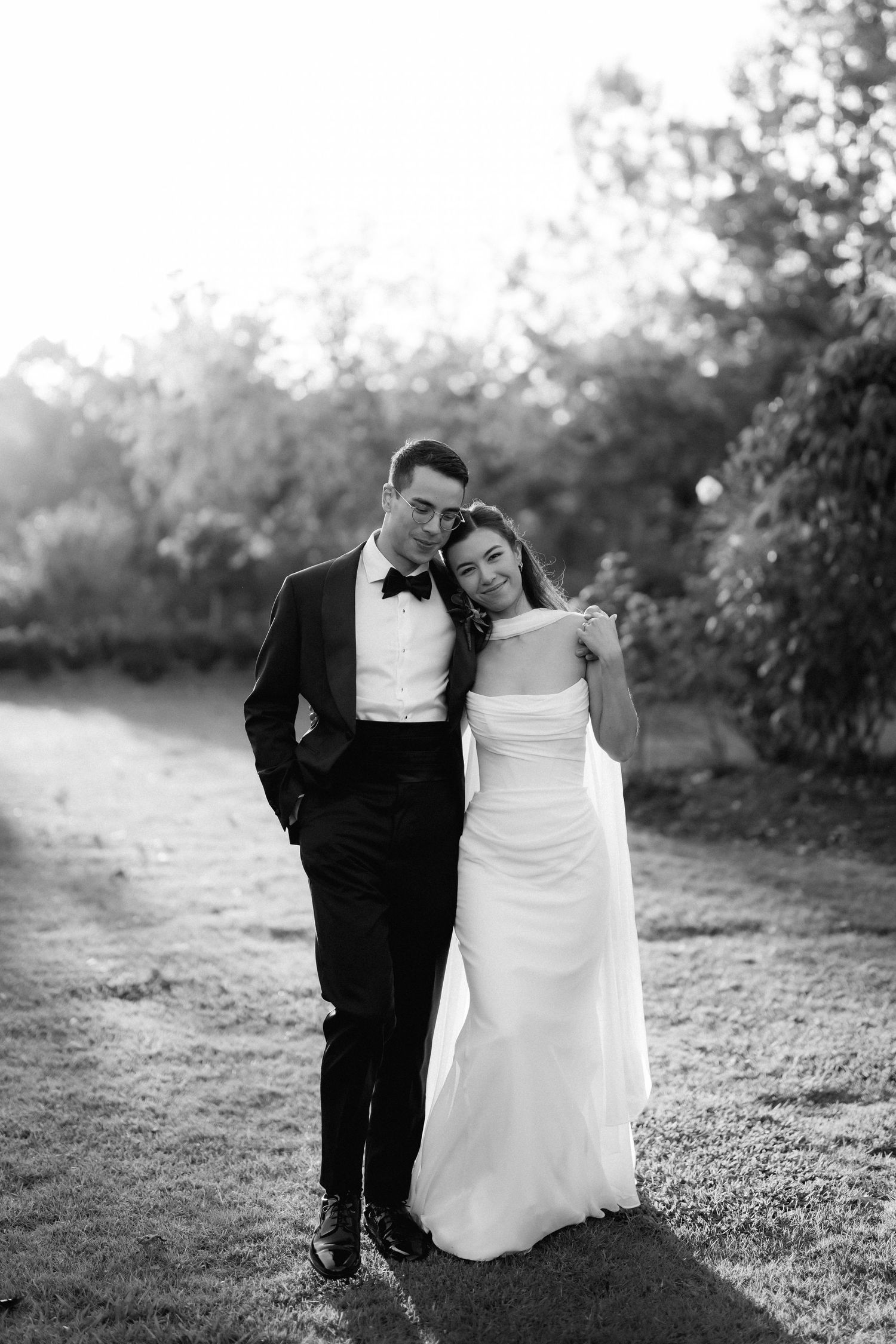 Black and white portrait of newlywed couple embracing outdoors, groom in tuxedo and bride in gown.