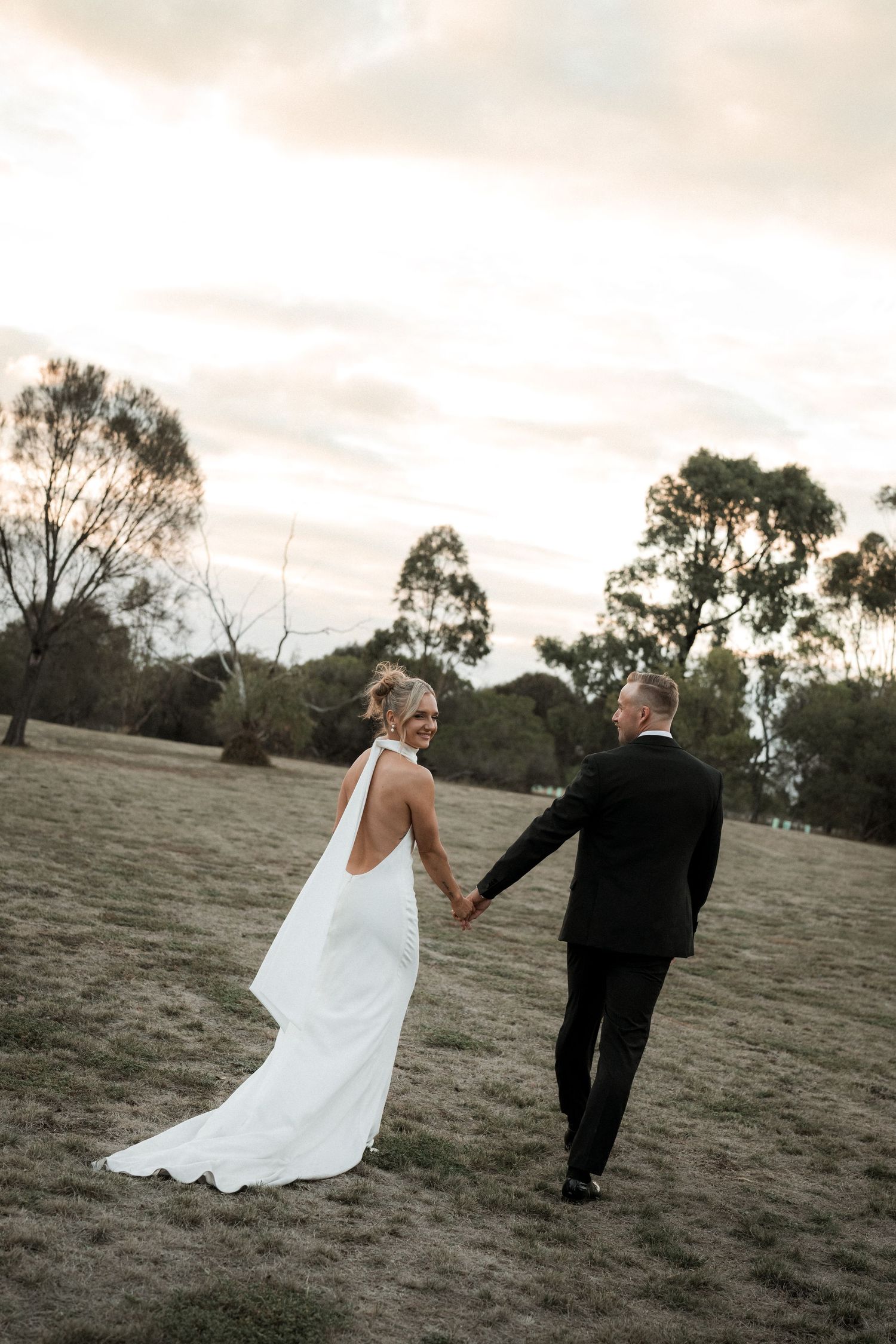 Newlywed couple walks hand in hand across a grassy field at sunset with eucalyptus trees in the background.