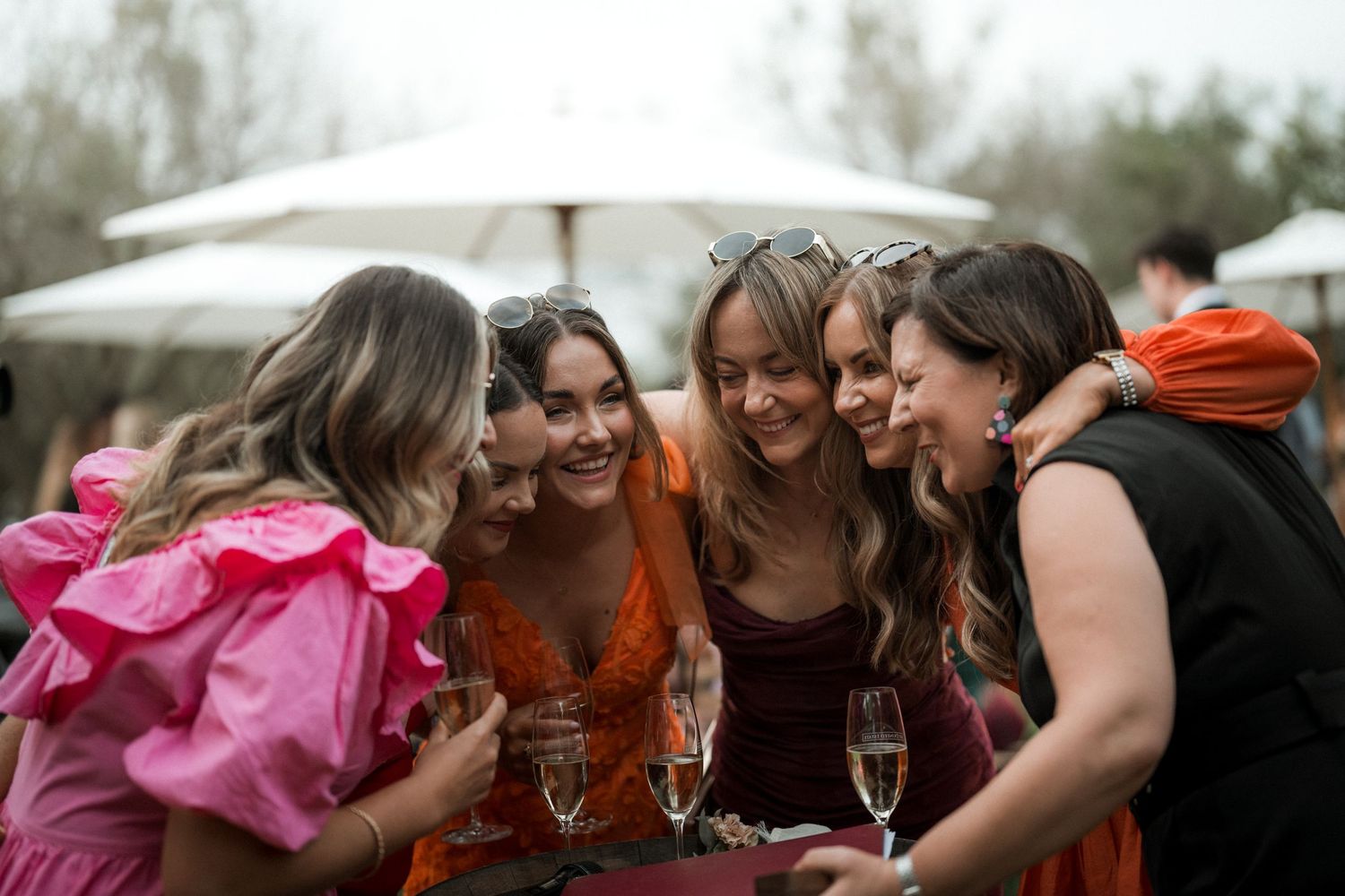 A group of friends huddle together laughing and sharing drinks under umbrellas at an outdoor gathering.