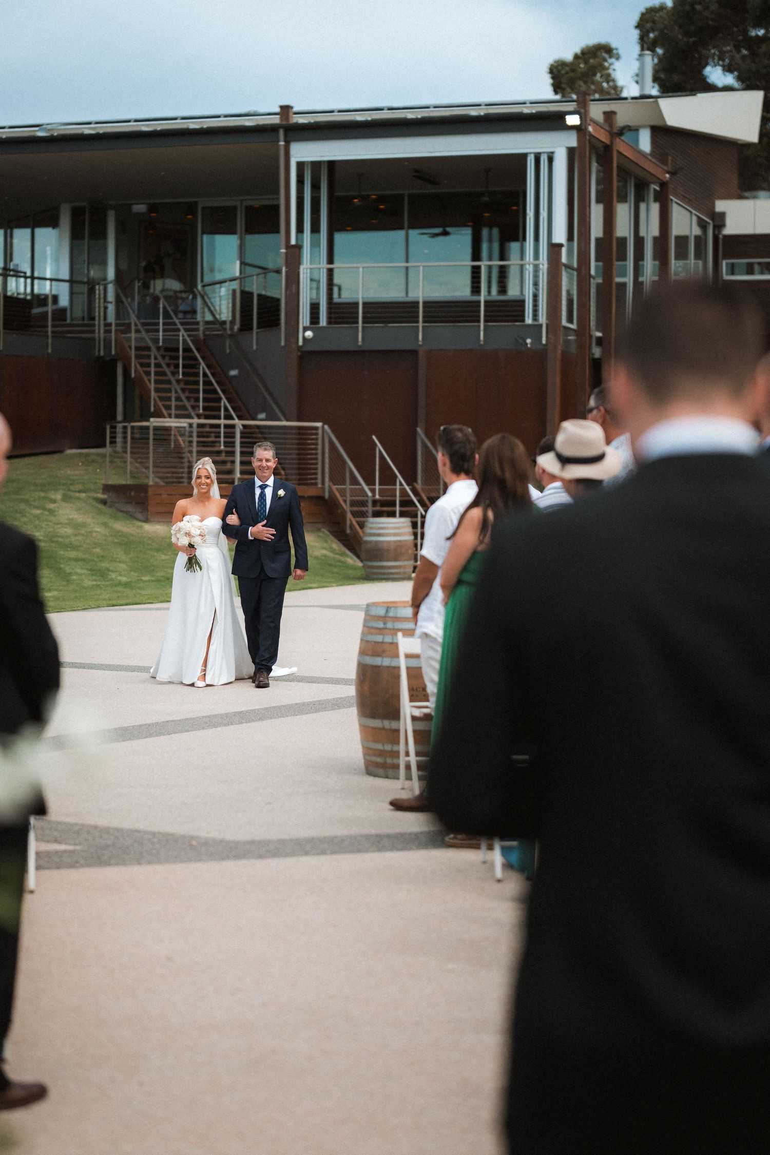 A wedding ceremony taking place outside a modern building with guests watching and a wine barrel as decor.