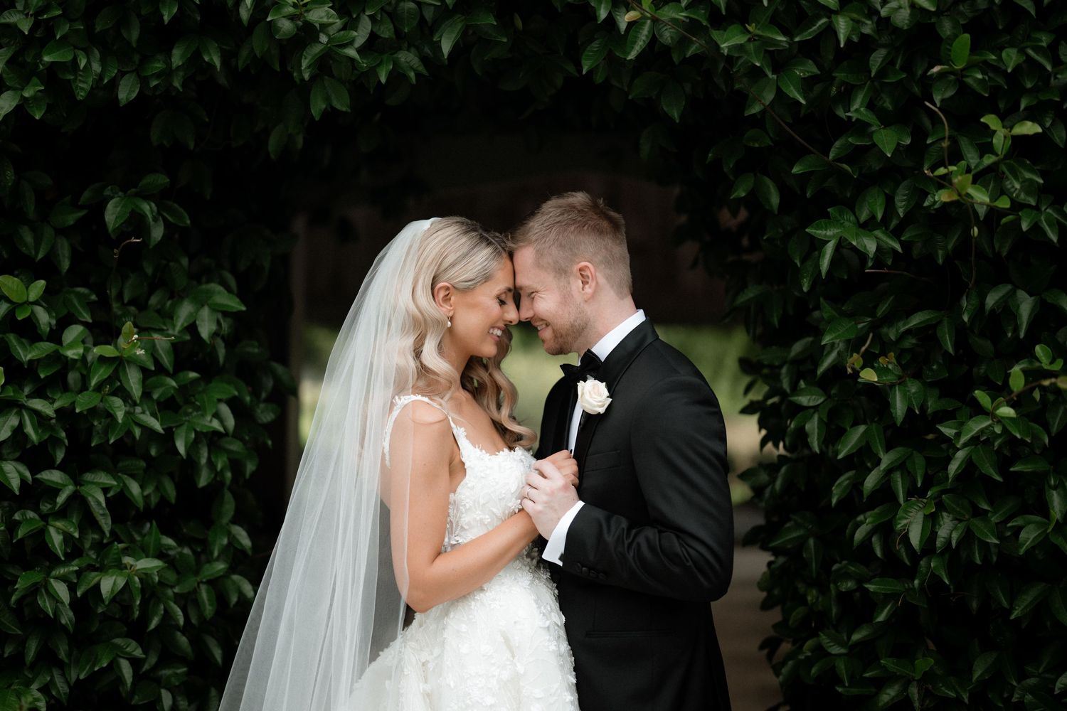 A romantic wedding couple shares an intimate moment against a backdrop of lush green foliage.