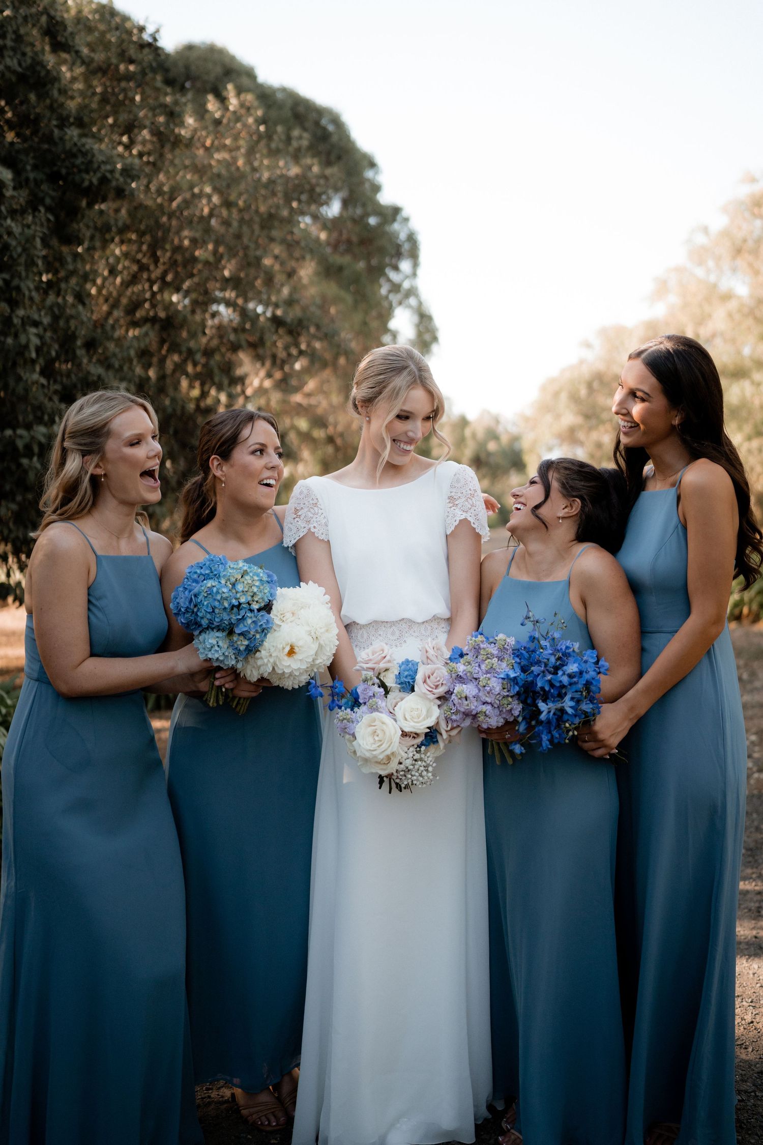Bridesmaids in dusty blue dresses hold blue and white bouquets while posing with bride at outdoor wedding.