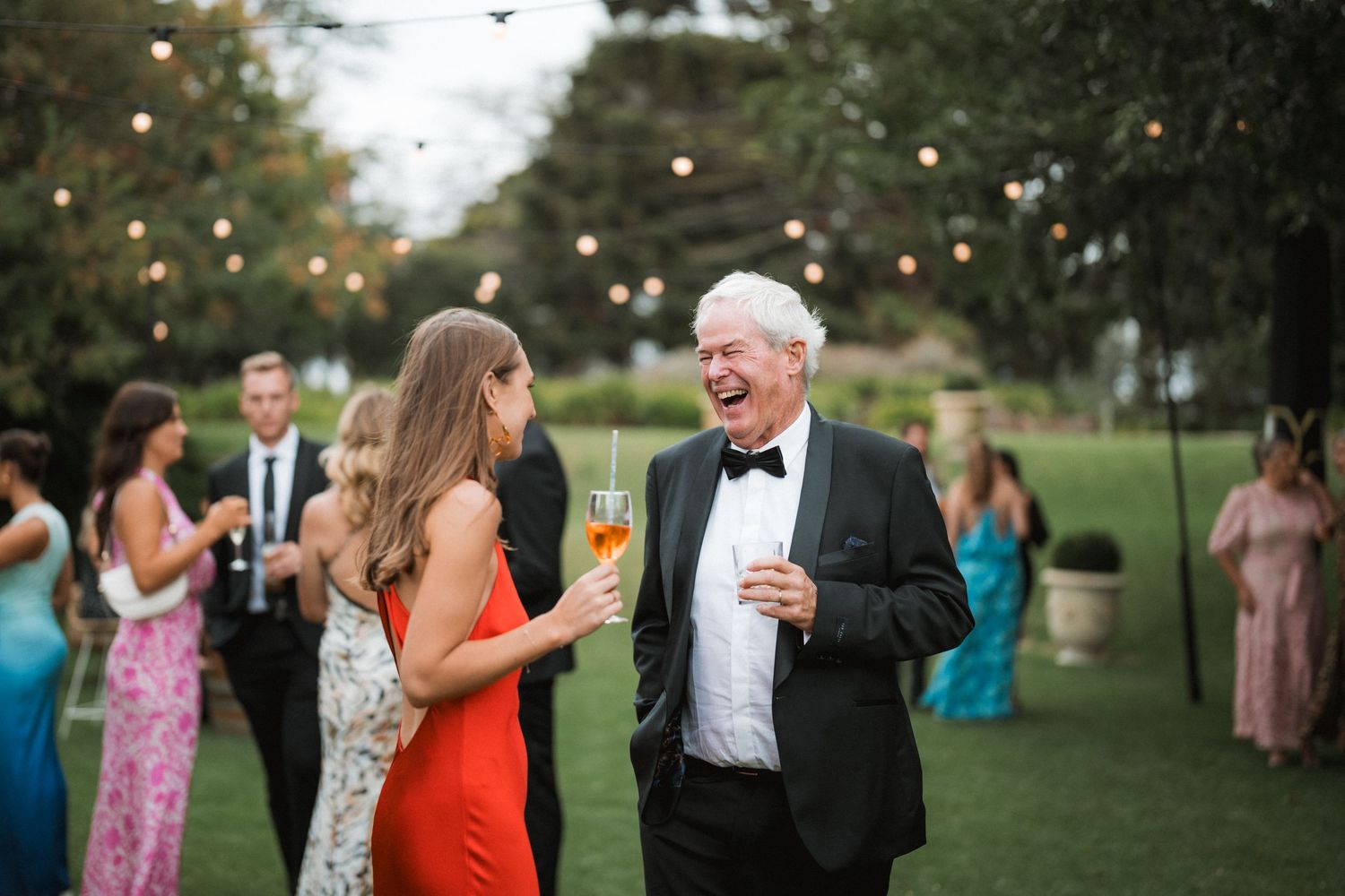 A lively outdoor wedding reception with people laughing and mingling on a grassy lawn under string lights at dusk.