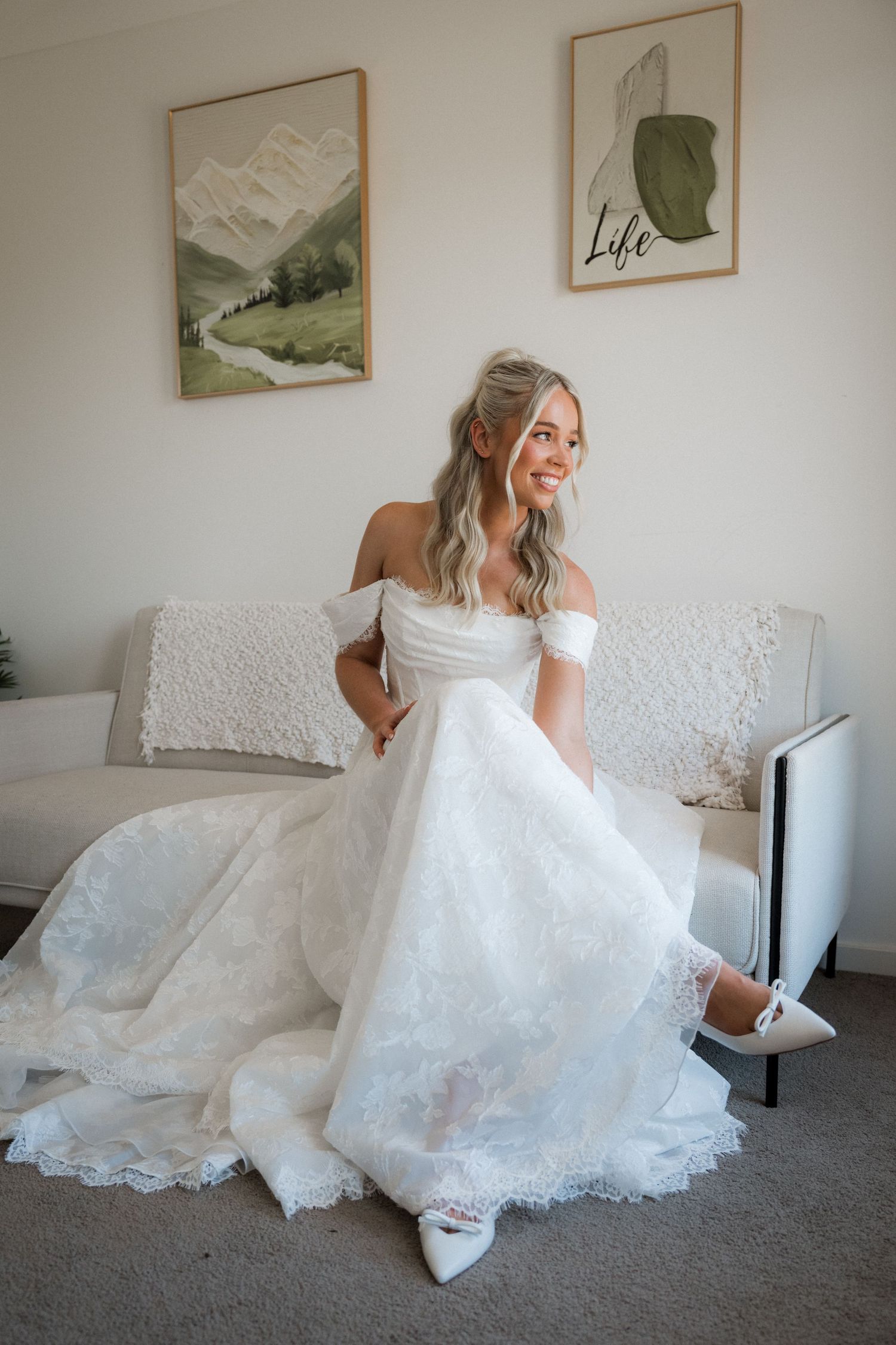 Smiling bride in off-shoulder lace wedding gown sitting on white sofa with art prints on wall behind her.