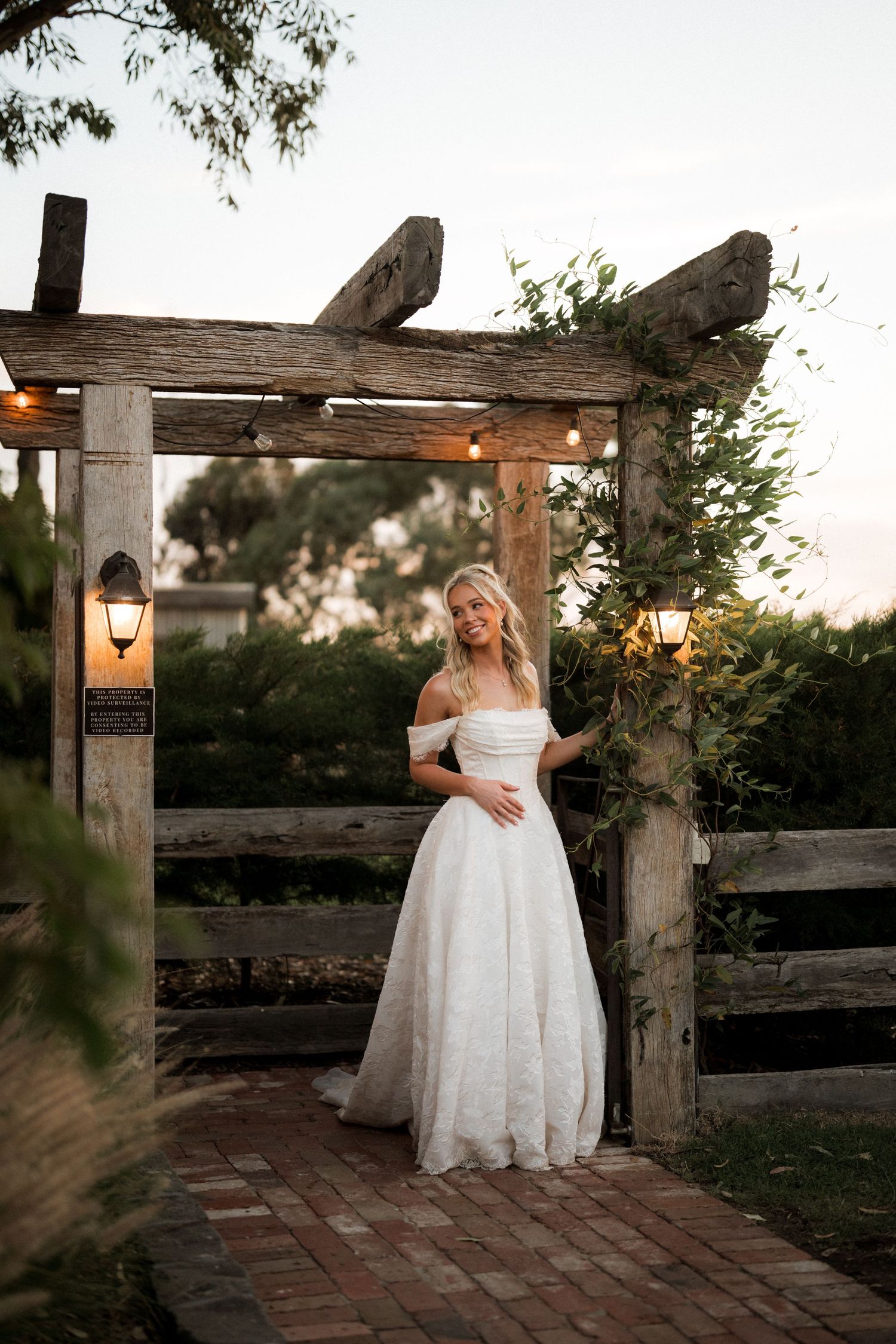 Smiling bride in off-shoulder lace gown standing under rustic wooden pergola at twilight.