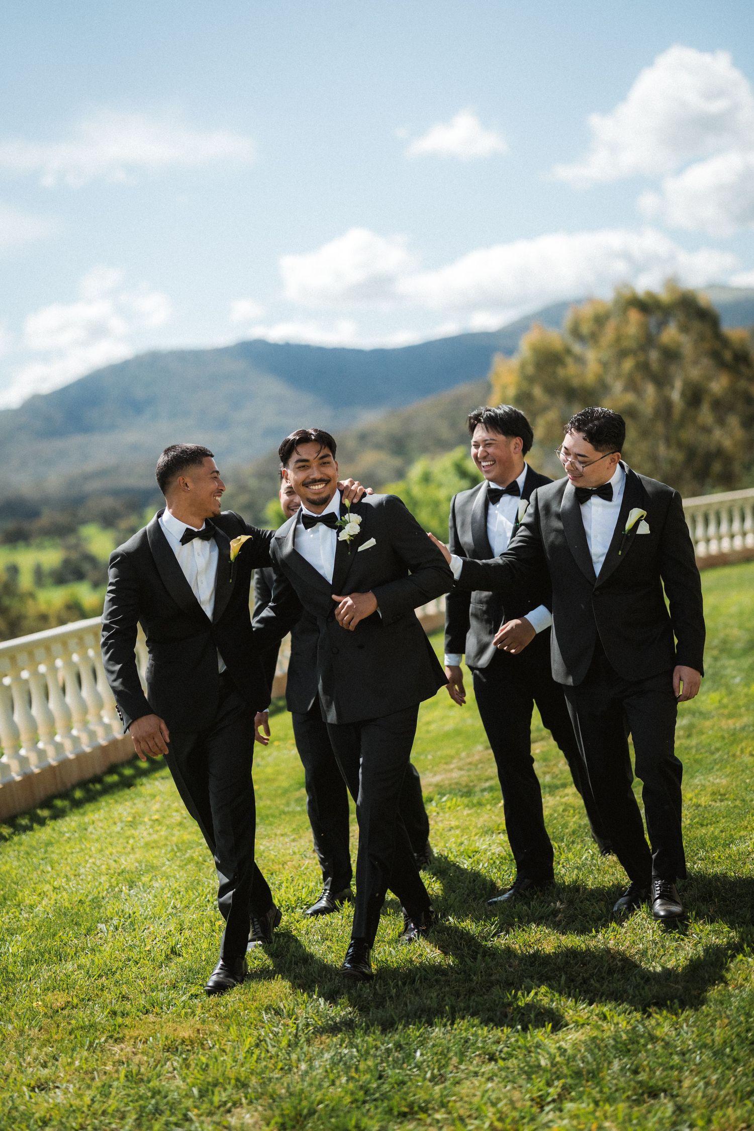 Five groomsmen in black tuxedos and bow ties walk and laugh together on a grassy lawn with mountain views at a wedding.