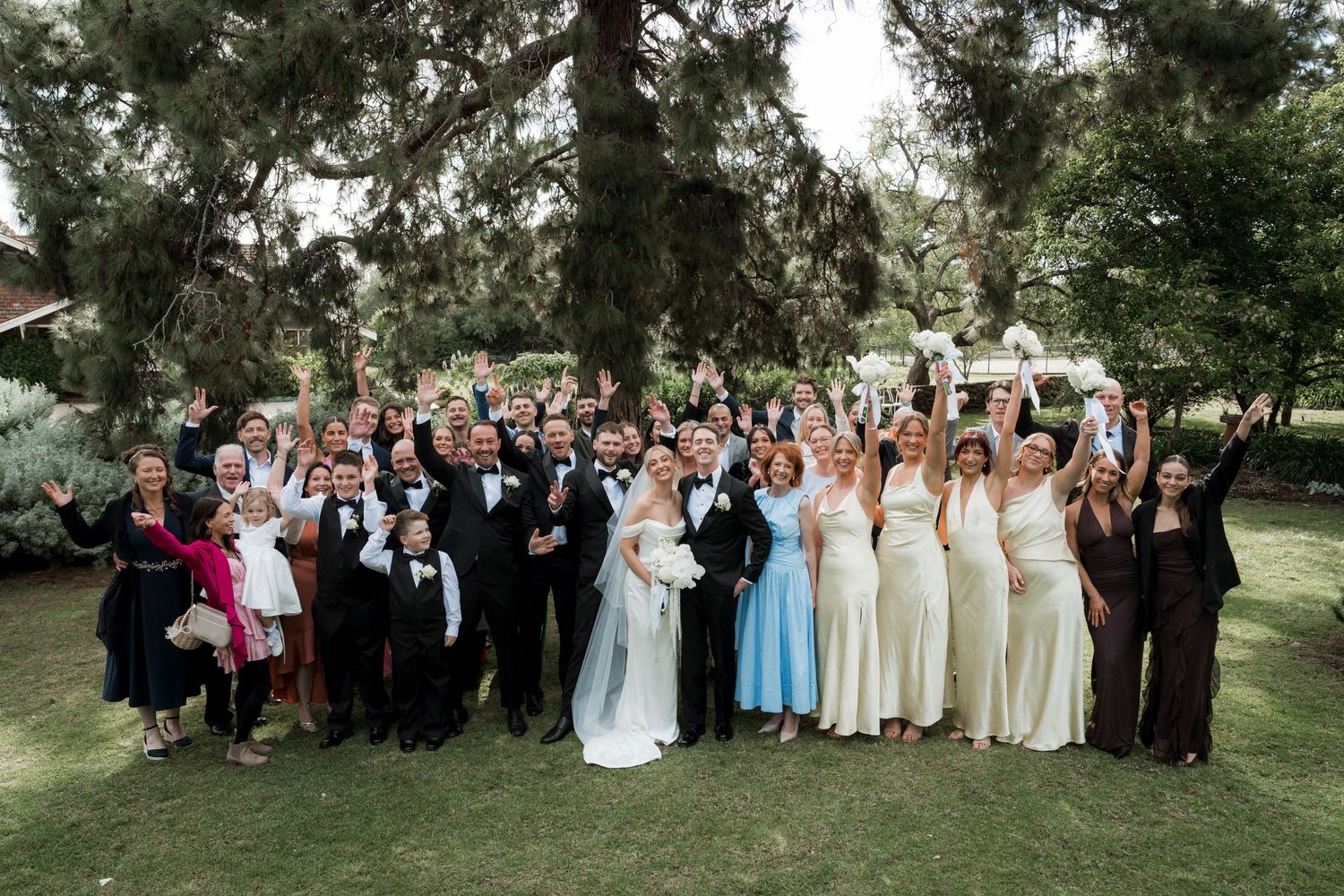 Large wedding party poses for a group photo outdoors with trees in the background.