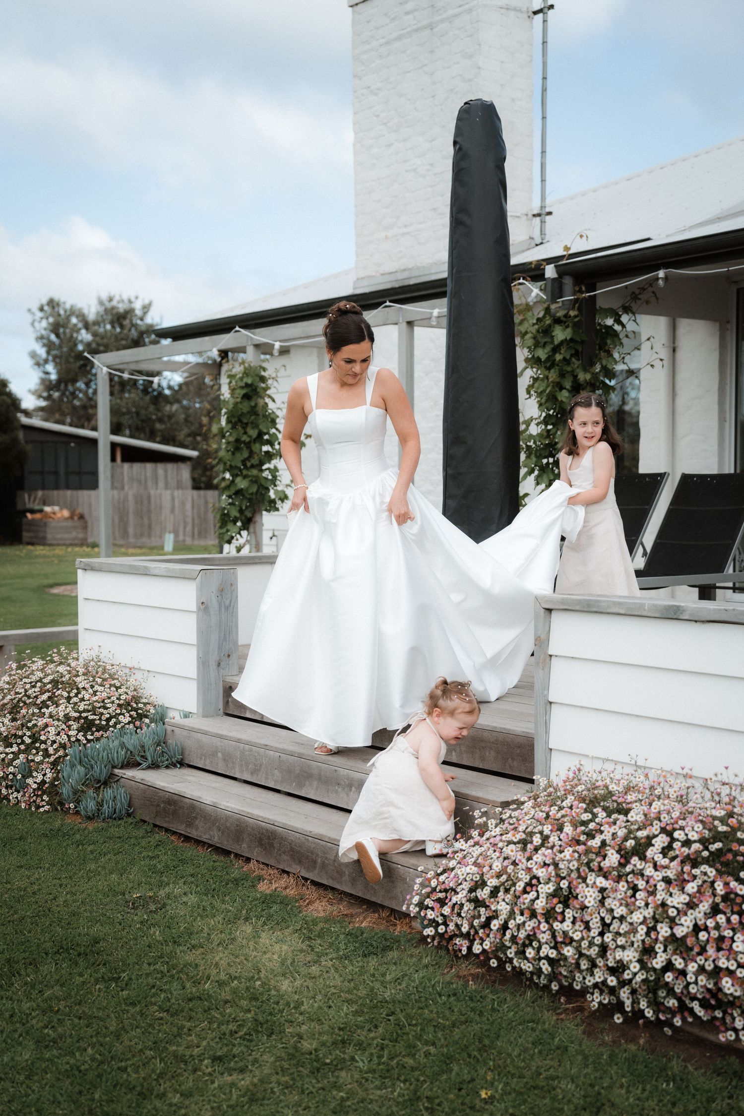 Bride in a tea-length wedding dress playfully poses by white railings with pink flowers in the garden.