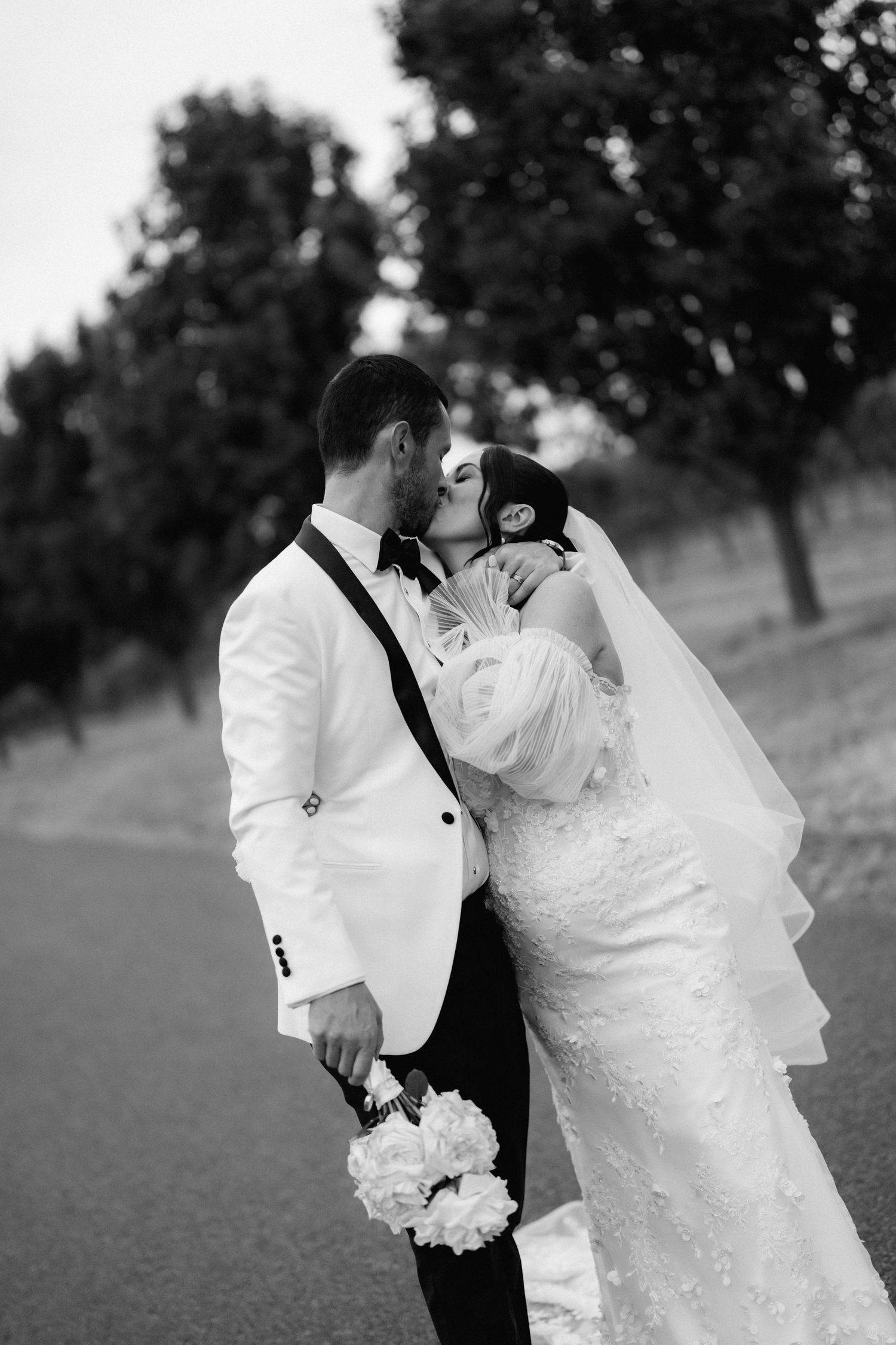Black and white wedding photo of a couple embracing on a tree-lined path with white bouquet.