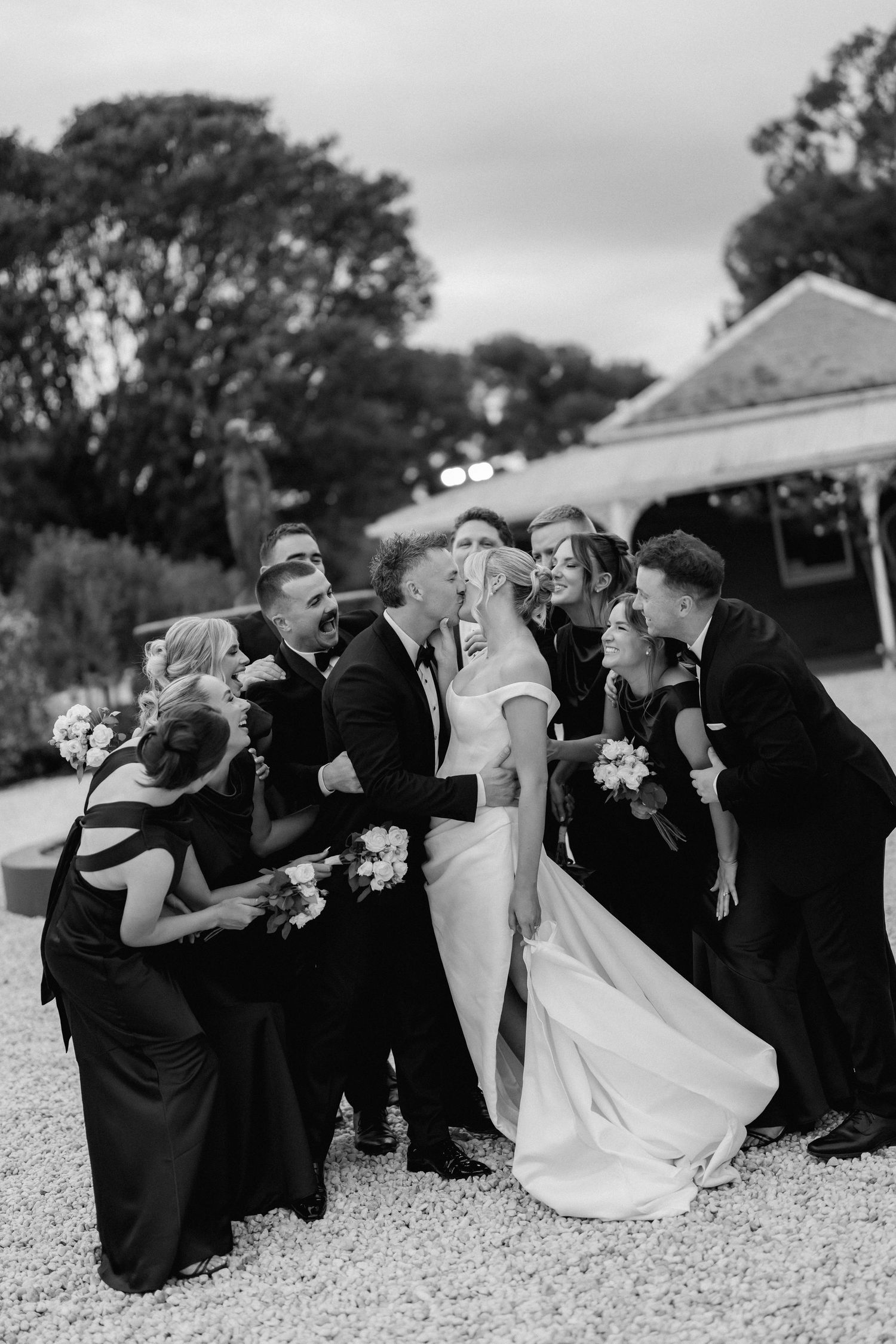 Wedding party in black attire performs synchronized dance move on gravel path in black and white photo series.