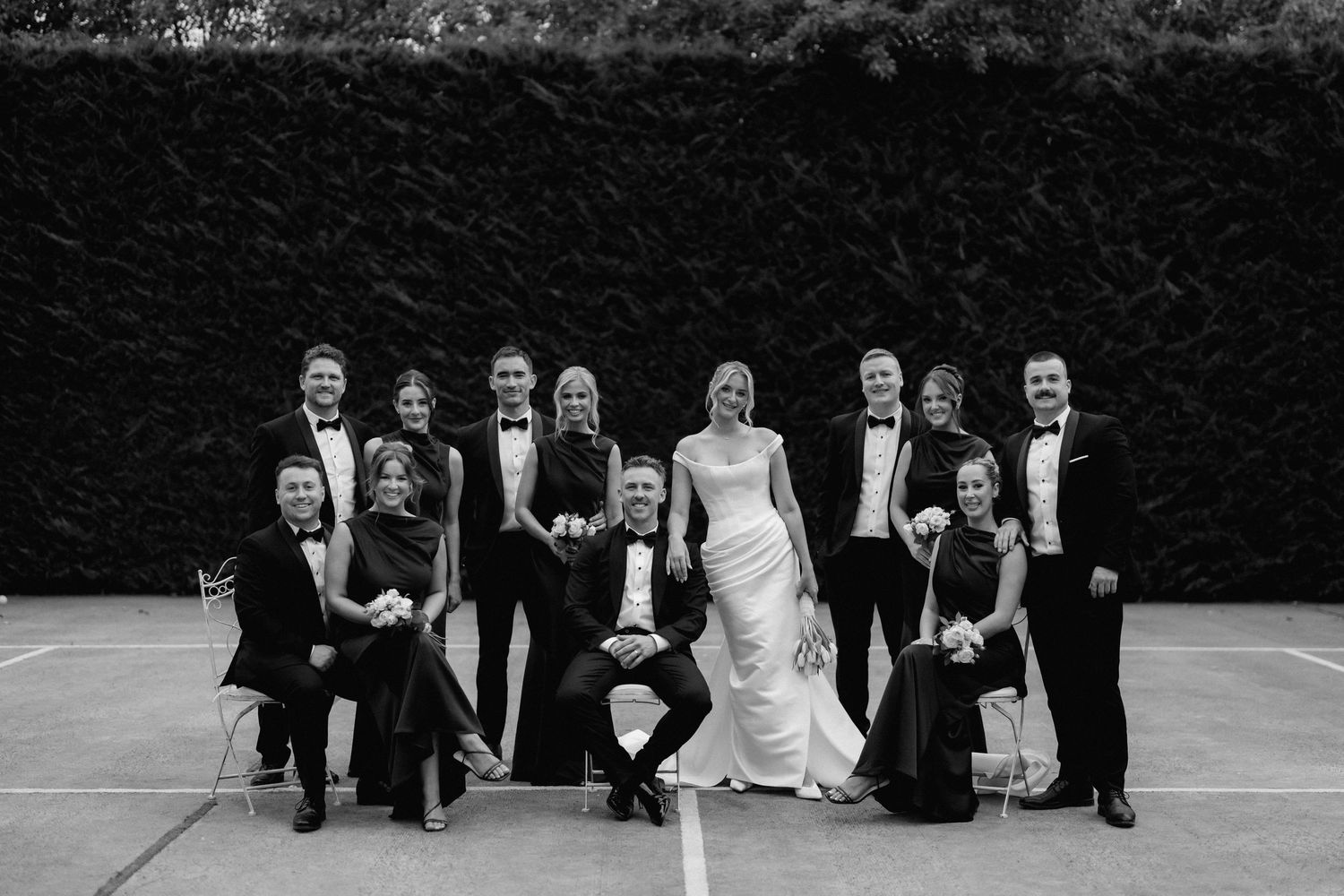 A wedding party in formal black and white attire poses together outdoors against a dark hedge backdrop.