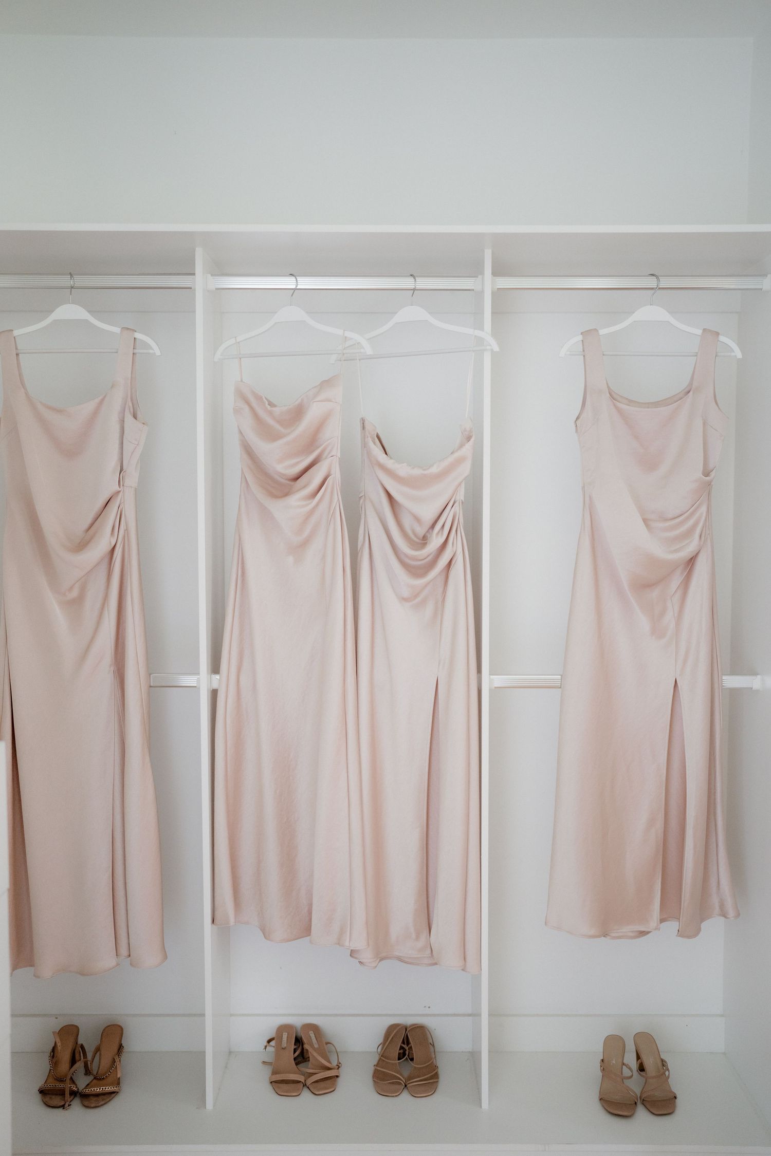 Blush pink bridesmaid dresses and nude heels arranged neatly on white hangers against a light wall.
