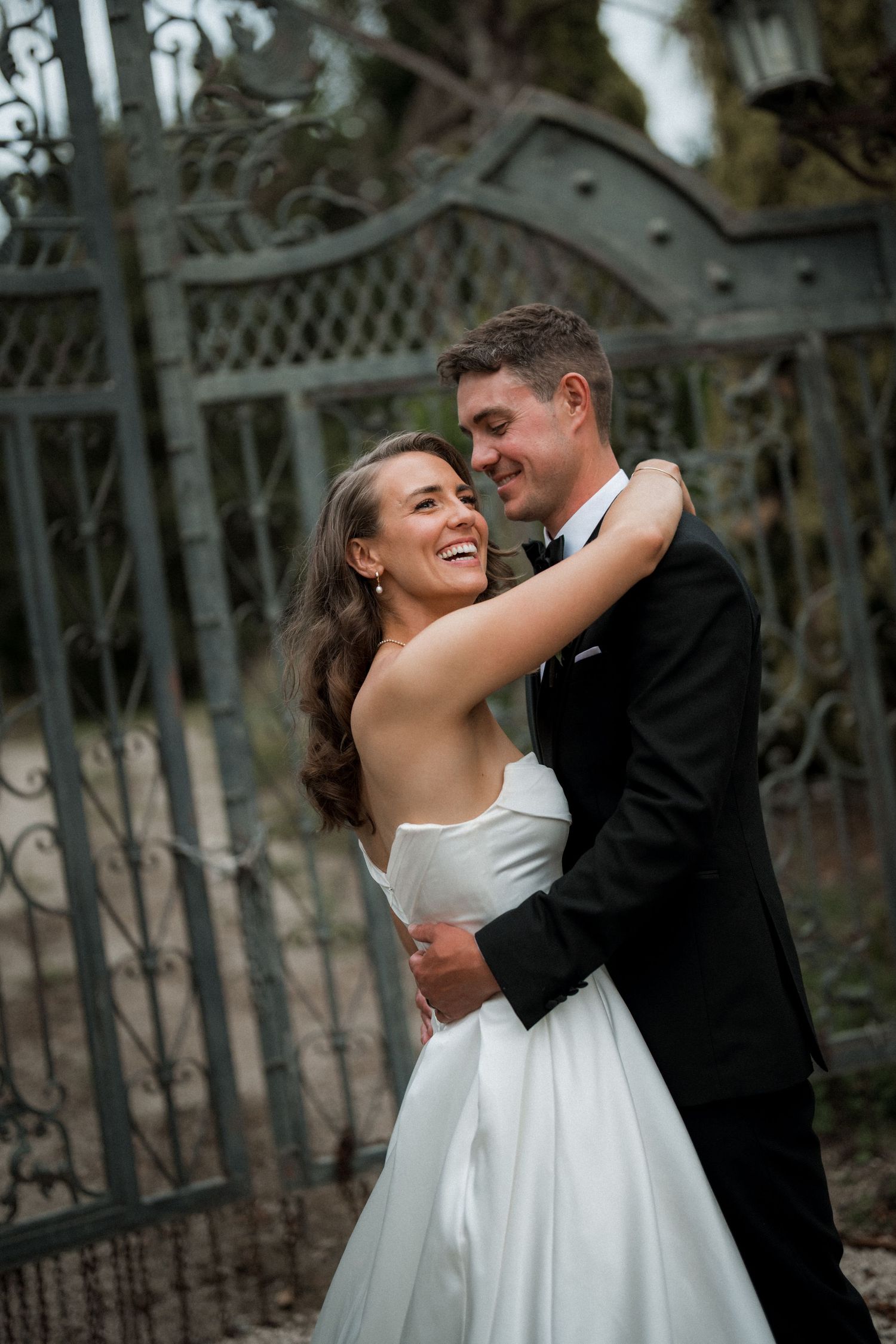 A romantic wedding couple embraces and laughs together in front of an ornate iron gate during their photo session.