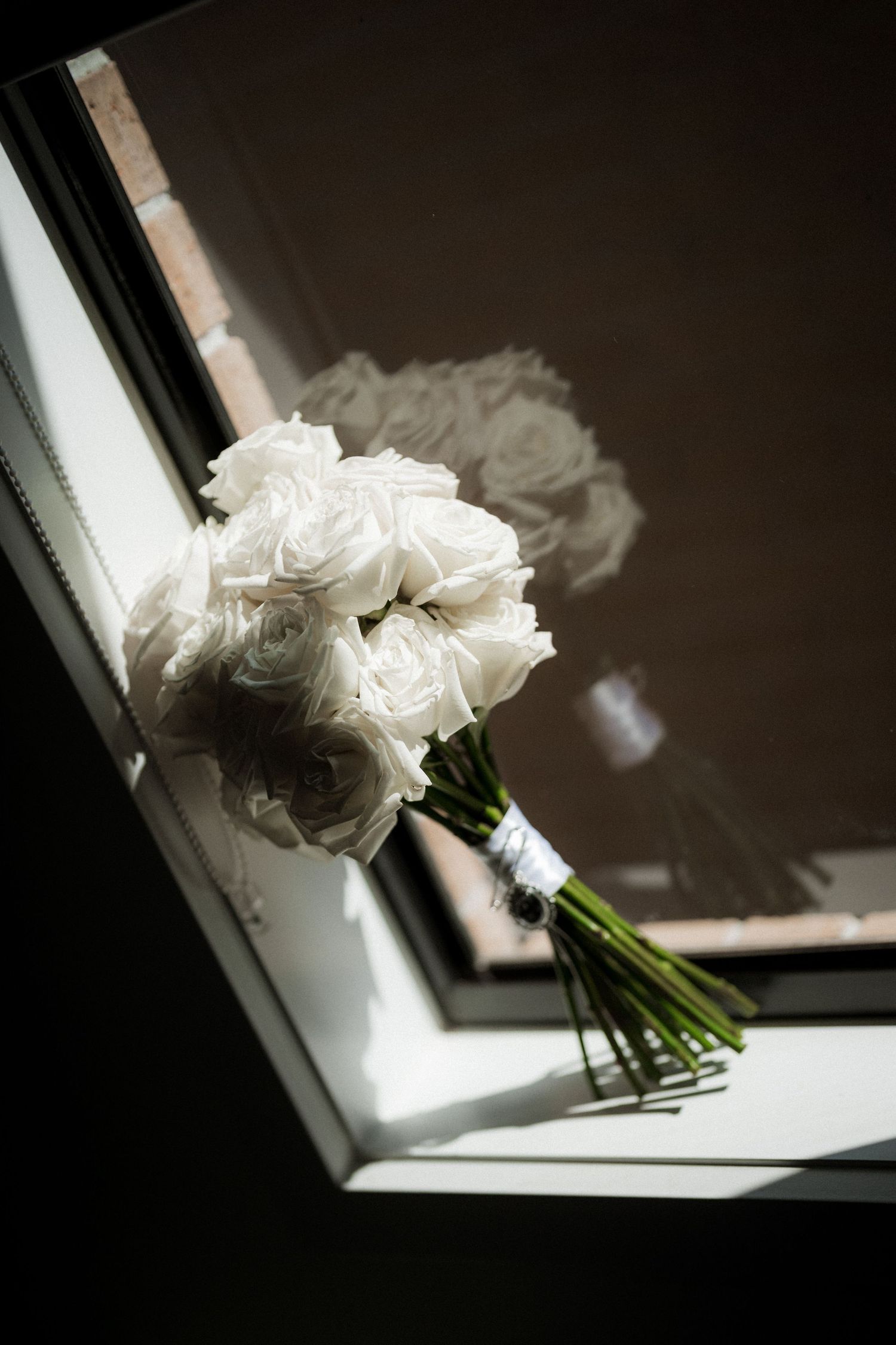 A white rose bouquet sits in dramatic lighting on a windowsill, casting soft shadows against a dark background.