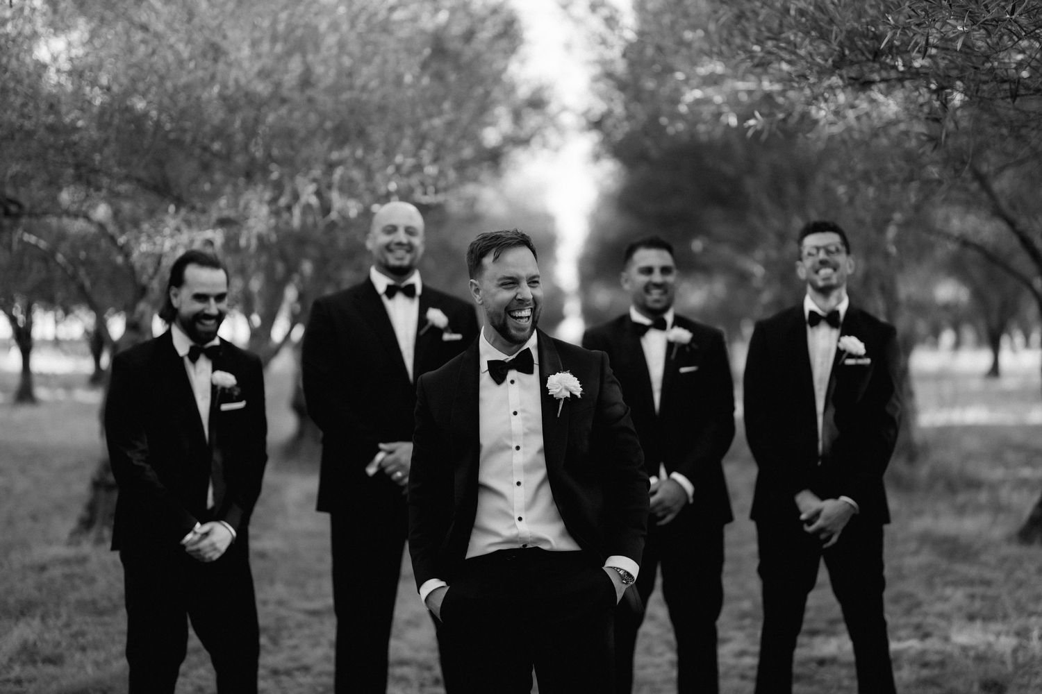 A group of groomsmen in black tuxedos stand together outdoors for a formal wedding photo.