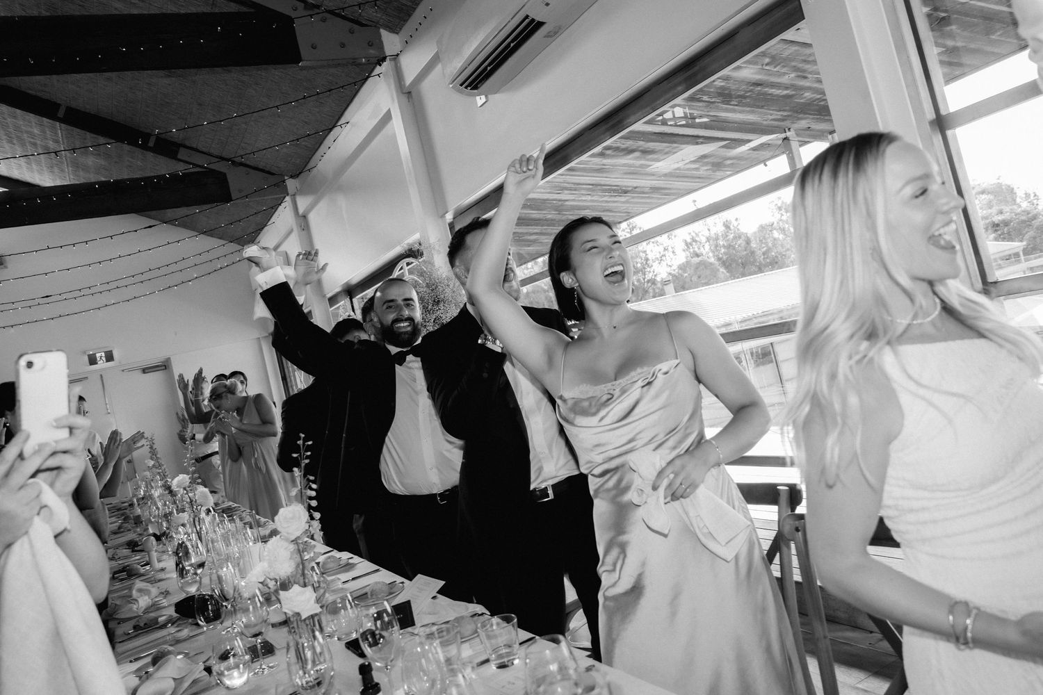 Wedding reception guests dancing and celebrating under a white tent ceiling with exposed beams.