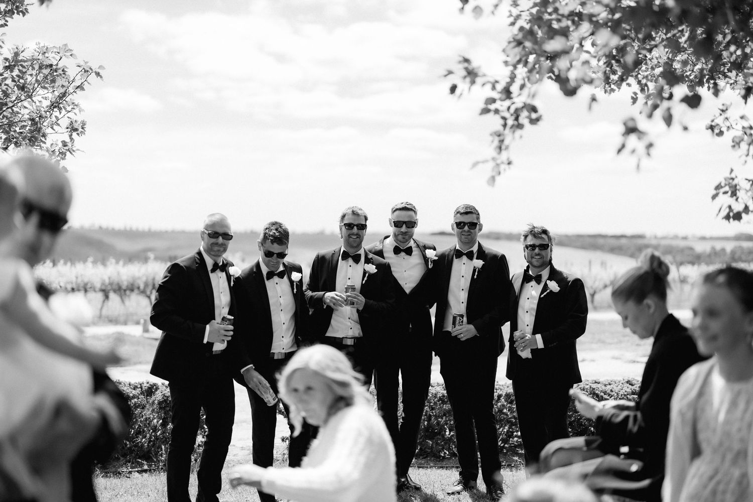 Wedding party in formal attire laughs together while walking outdoors on a sunny day.
