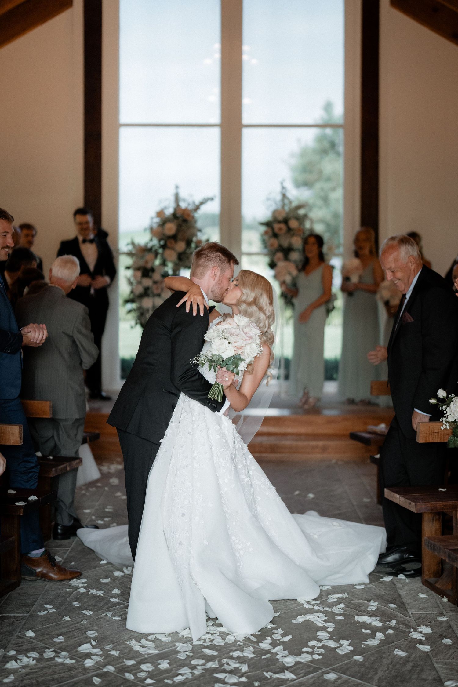 A romantic wedding kiss in a chapel with large windows, bridesmaids in blue dresses, and rose petals scattered on the floor.